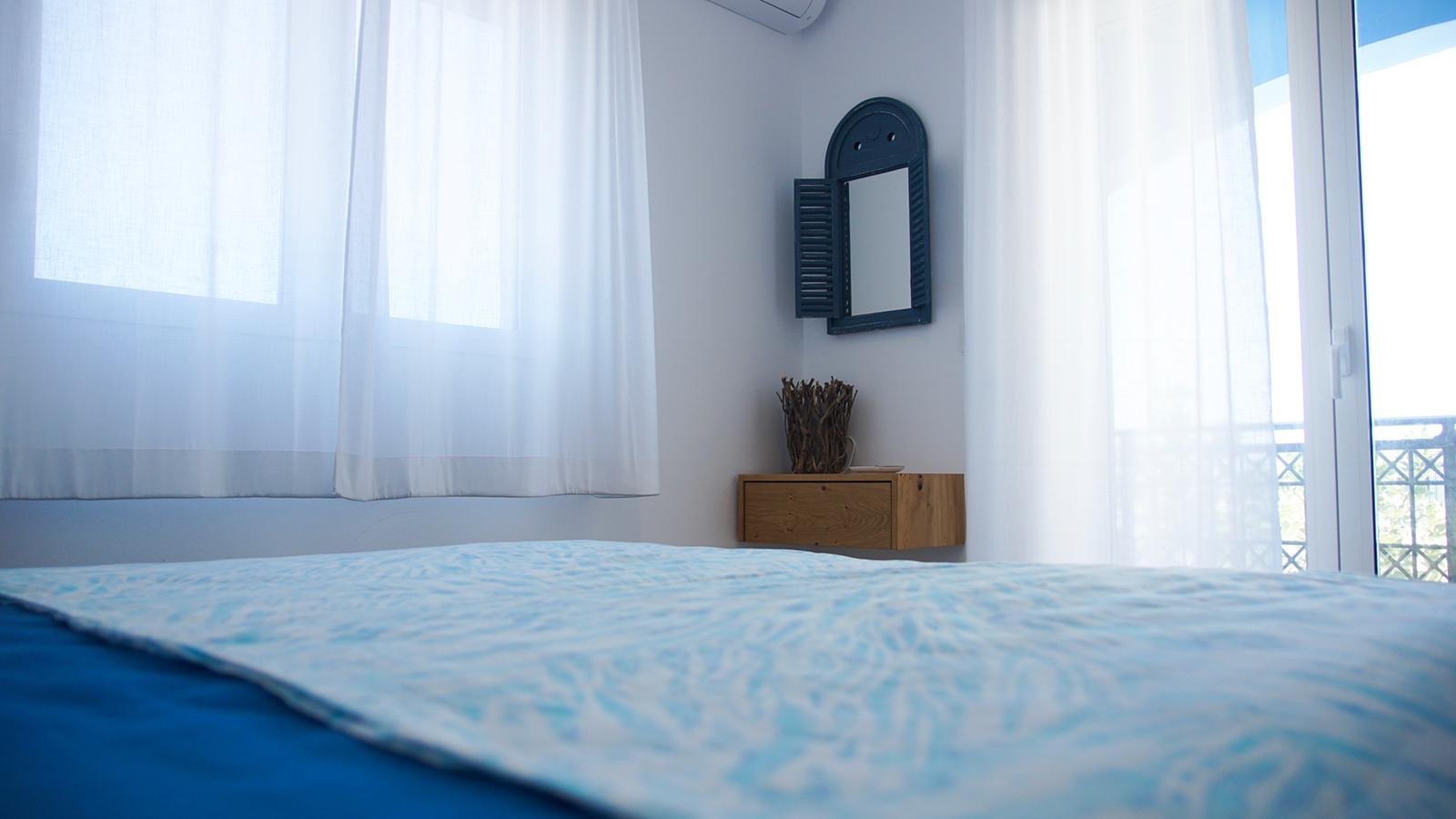 Ein Schlafzimmer mit weißen Vorhängen, einem kleinen Holznachtisch, einer Dekorationsbox und einem Wandspiegel.