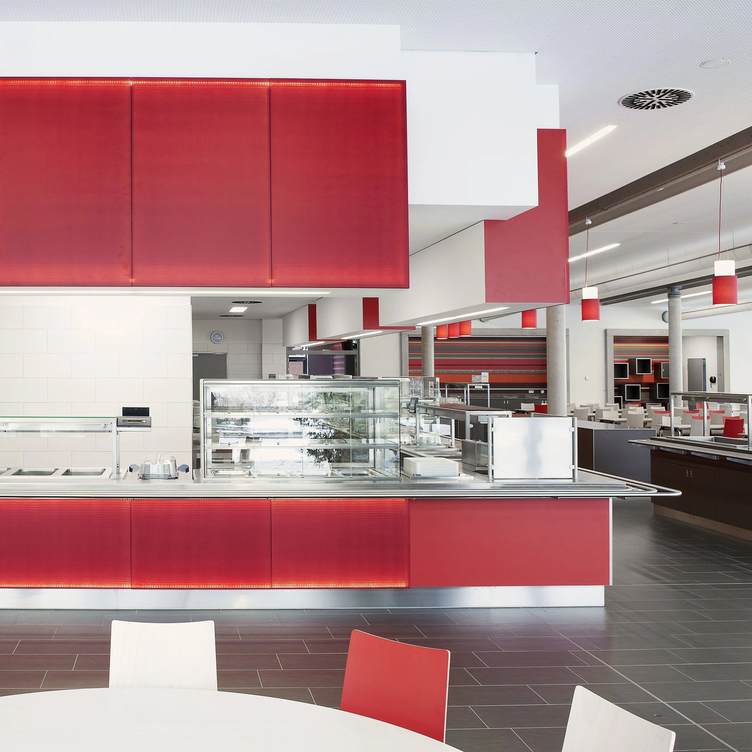 Innenansicht eines modernen Cafeterias oder Schnellrestaurants mit rotem und weißem Design, Sitzplätzen im Hintergrund, und einem Verkaufsbereich mit Schalen und Ablagen im Vordergrund.