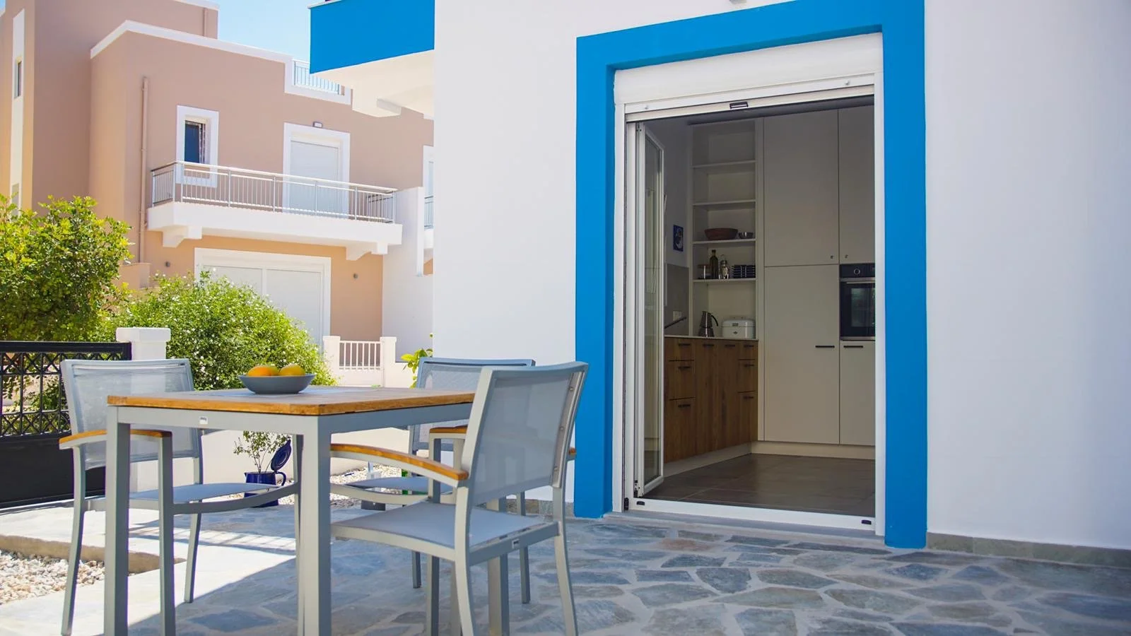 Blick auf eine Terrasse mit einem Esstisch und Stühlen, offener Blick auf eine moderne Küche, im Hintergrund ausgebaute Mehrfamilienhäuser.