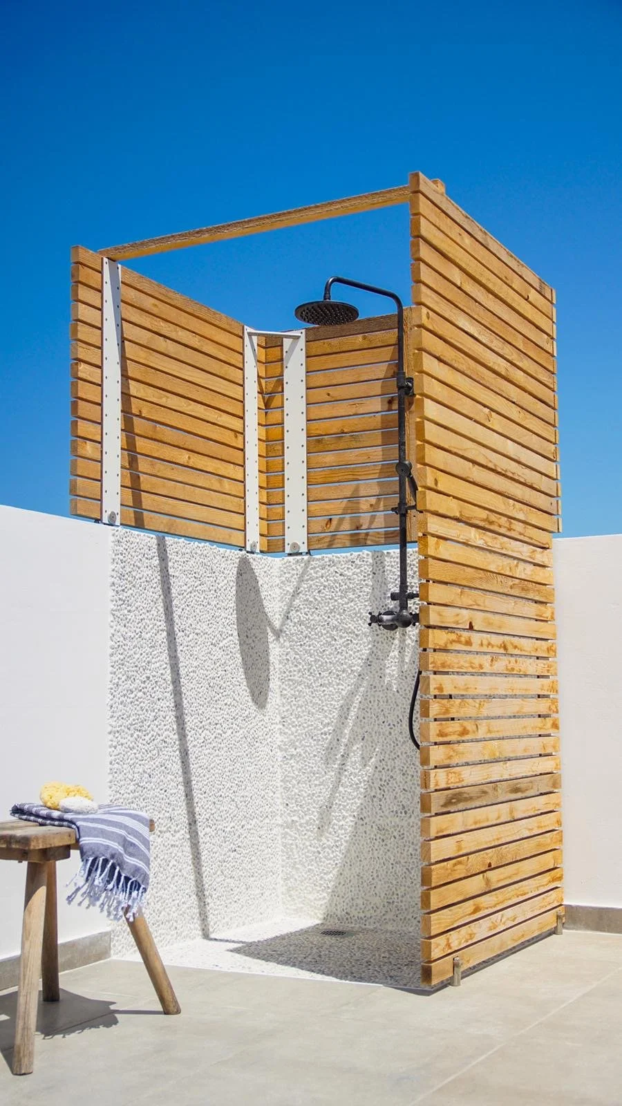Außendusche mit Holzwand und einem schwarzen Duschkopf, auf einer weißen Wand mit Kieselstein-Struktur, im Freien, bei blauem Himmel.