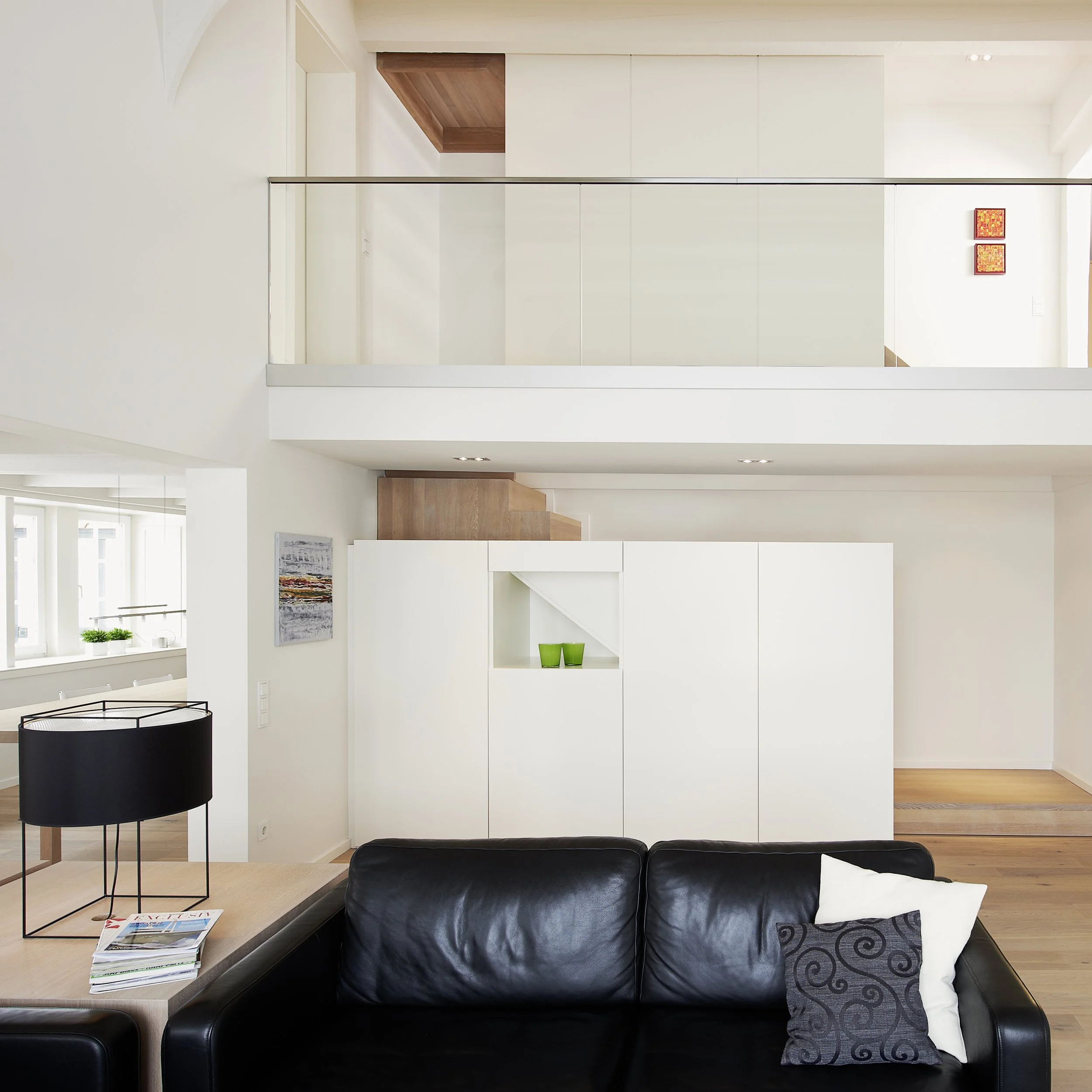 Modernes, minimalistisches Wohnzimmer mit schwarzen Ledersofa, weißen Wänden, Wandbild, Lampen und Treppe in Holzoptik, obere Etage mit Mezzanin