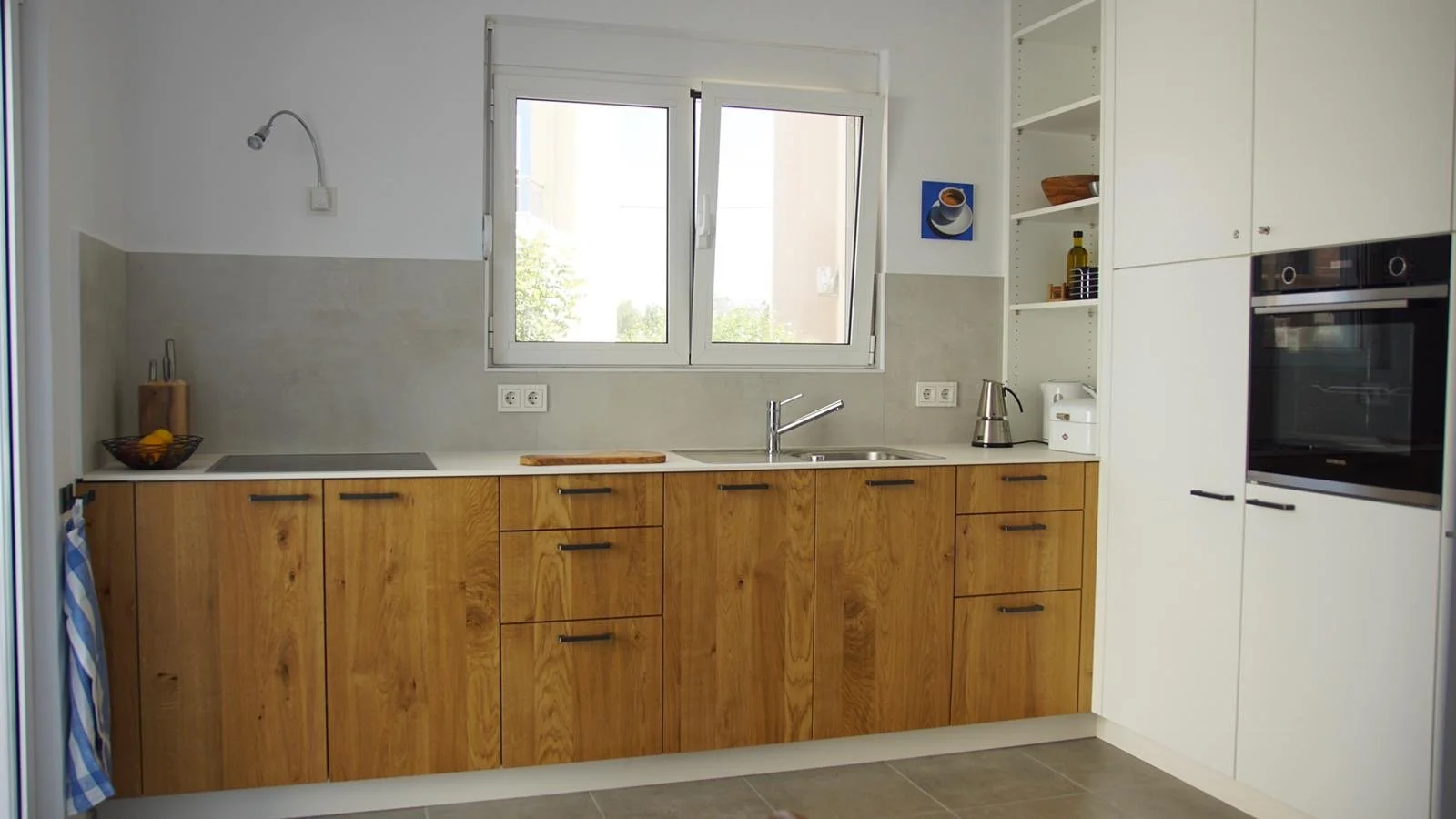 Moderne Küchenzeile mit Holzkabelfronten und weißen oberen Schränken, Fenster über der Spüle, elektrische Kochplatte, Wasserkocher und Geschirr auf den Regalen.