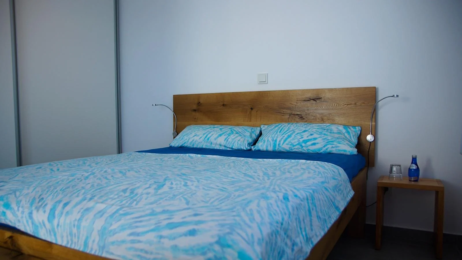 Ein Schlafzimmer mit einem Bett, Bettwäsche in Blau- und Weißtönen, zwei Kissen, Nachttisch mit Wasserflasche und Glas, und einem weißen Wandhintergrund.