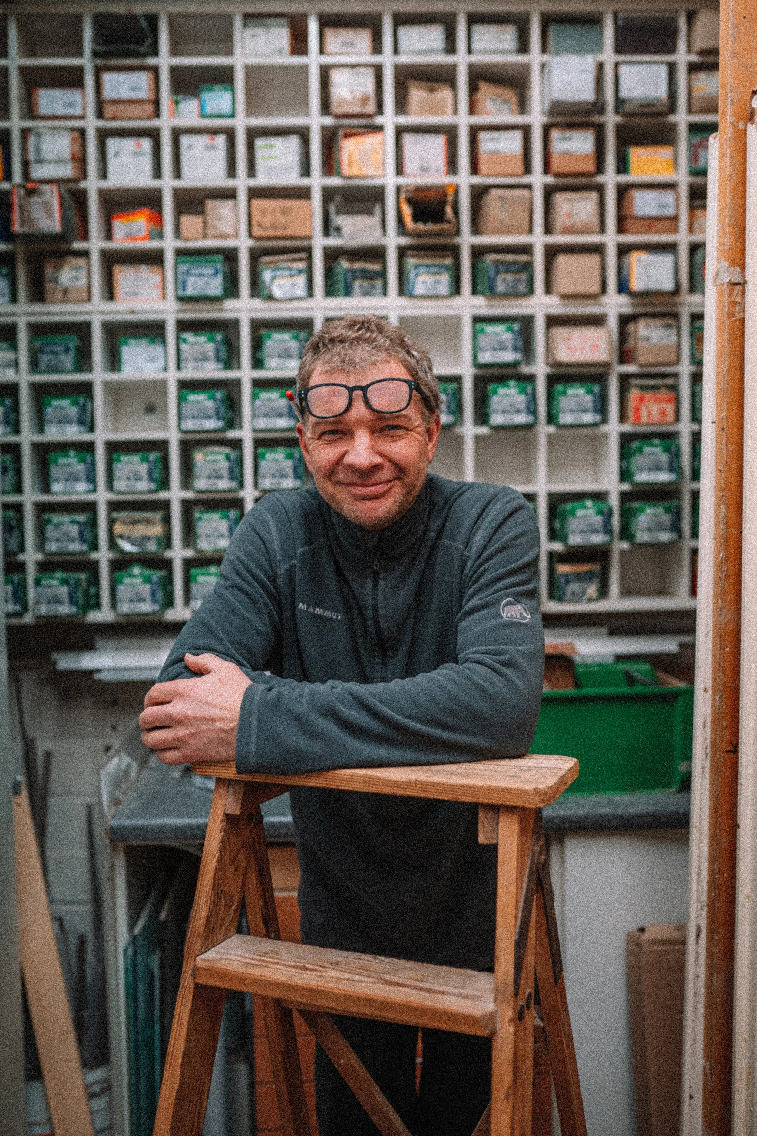 Ein Mann lächelt und sitzt in einem Workshopraum vor einem Holztrecker, im Hintergrund sind mehrere Fächer mit kleinen Kartons. Er trägt eine dunkle Jacke und eine Brille auf der Stirn.