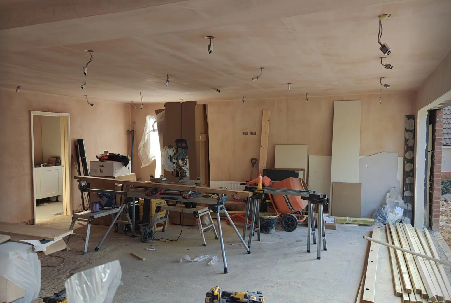 A room under renovation with construction tools, wood pieces, and plastering work in progress. Multiple workbenches and construction equipment are present.