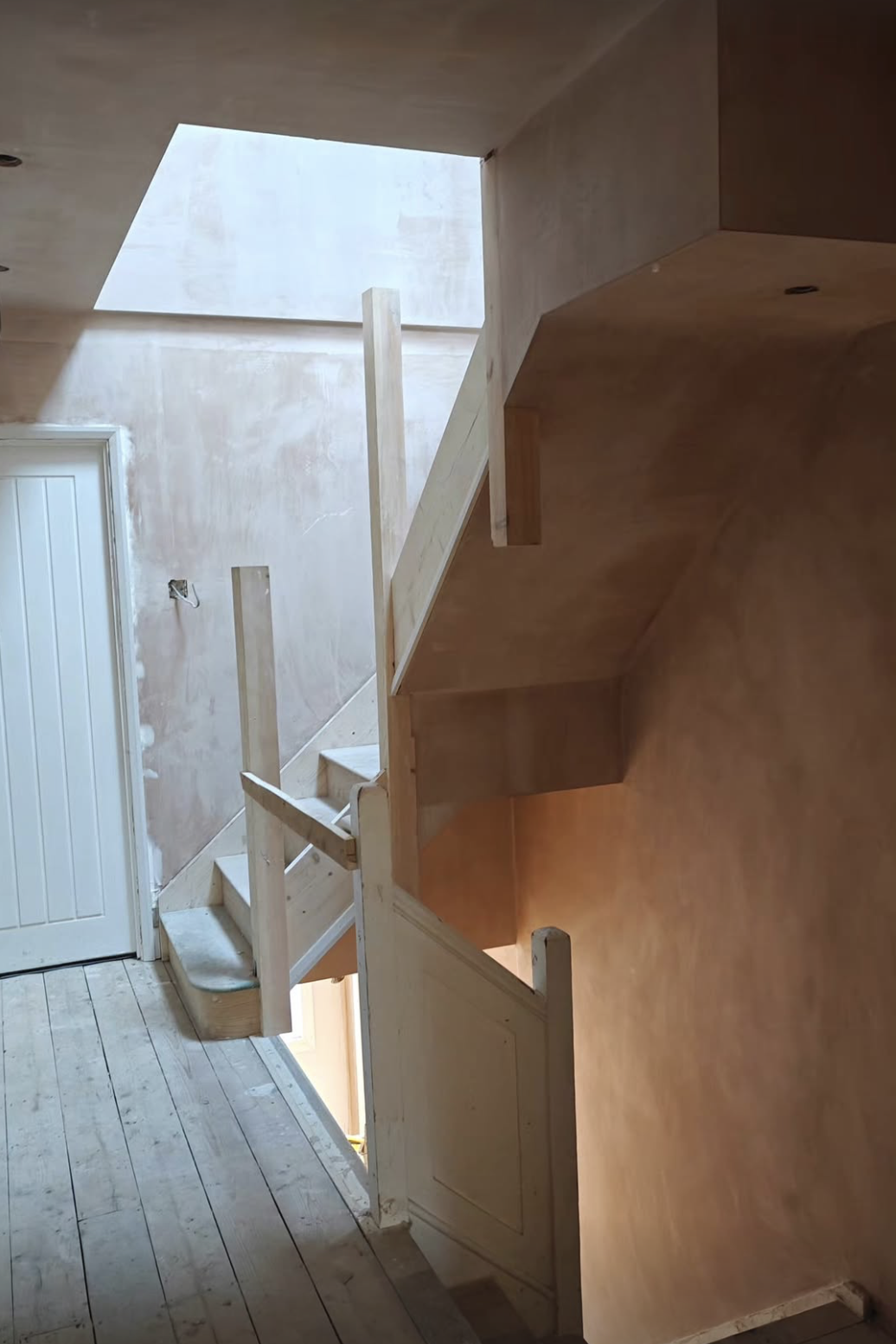 Photo of a house staircase under construction, featuring unfinished wood stairs, handrails, and plastered walls with a skylight overhead.