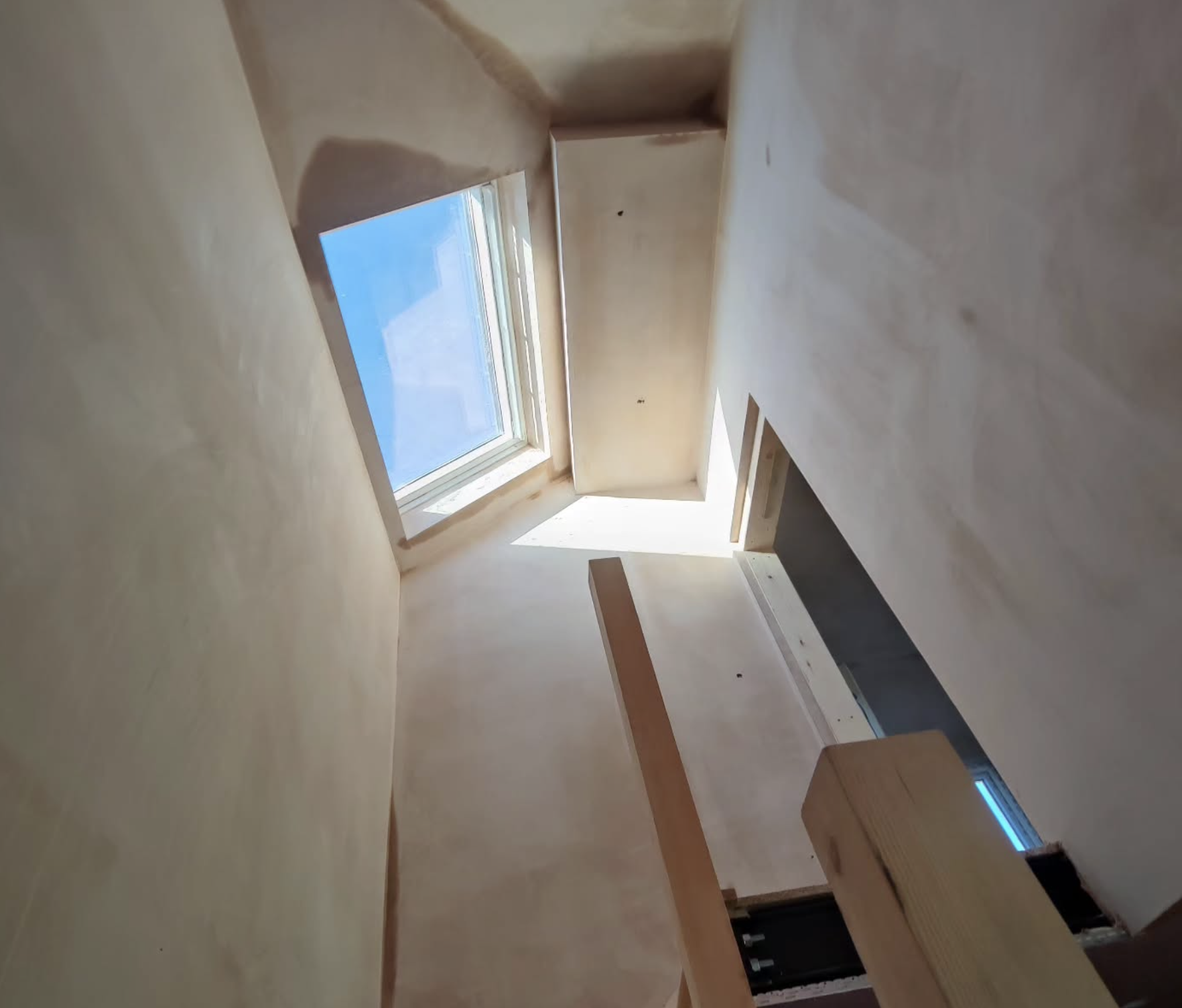 View from inside an attic with a skylight window showing a blue sky. The wooden structure, including freshly plastered wall, beams and frame, is visible.