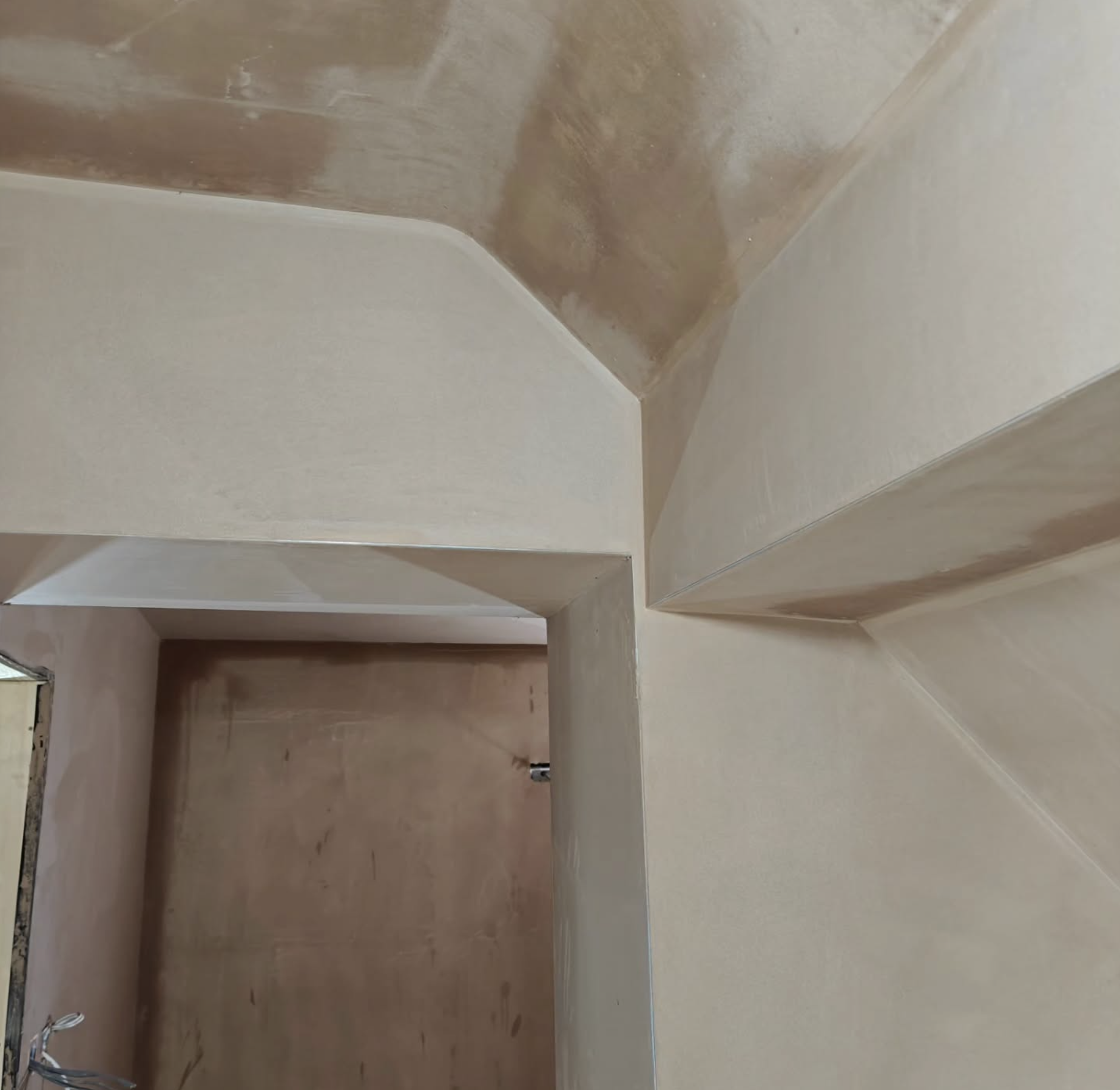 Interior space with freshly plastered walls, showing a corner with angular design elements and a doorway leading to another room.