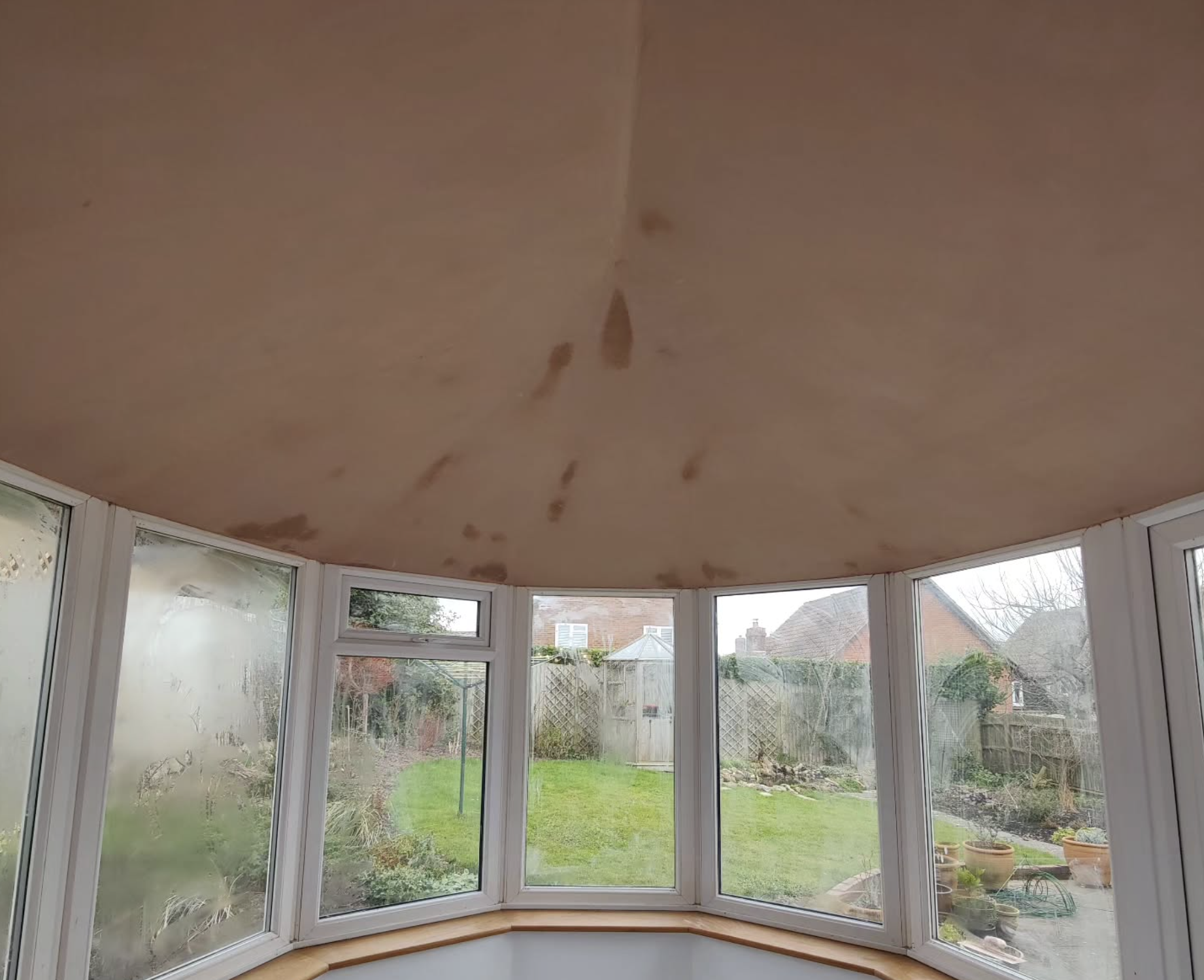 Ceiling with water stains in a sunroom with glass windows and a view of a backyard garden.