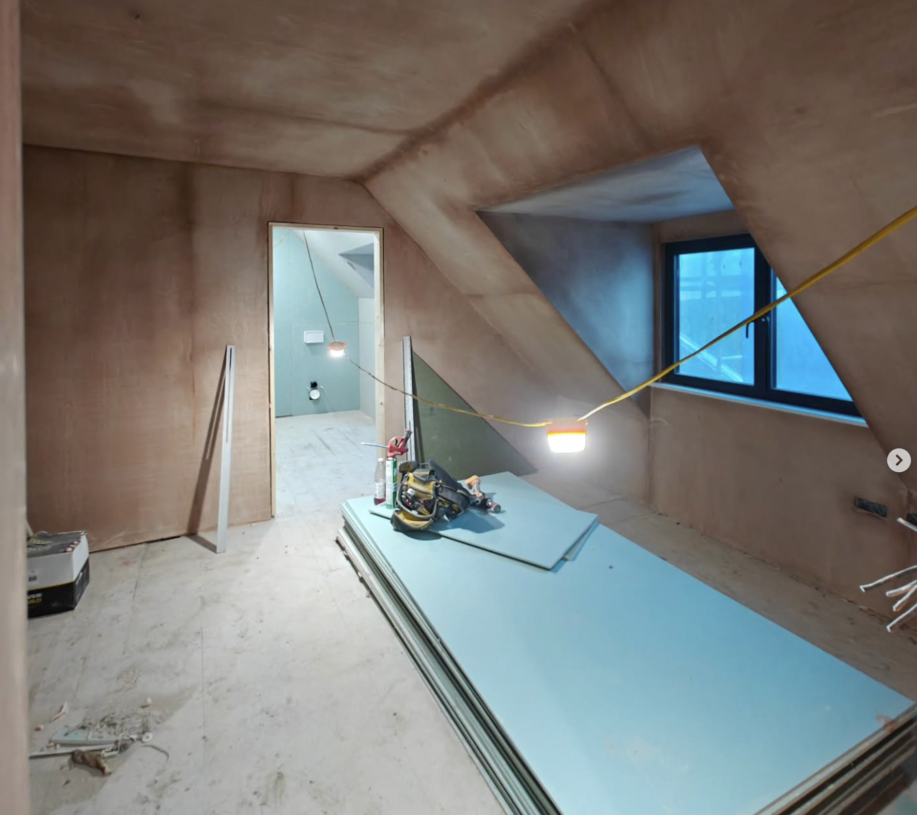 Construction site of an attic room with wooden walls, blue window, and construction tools and materials on the floor.
