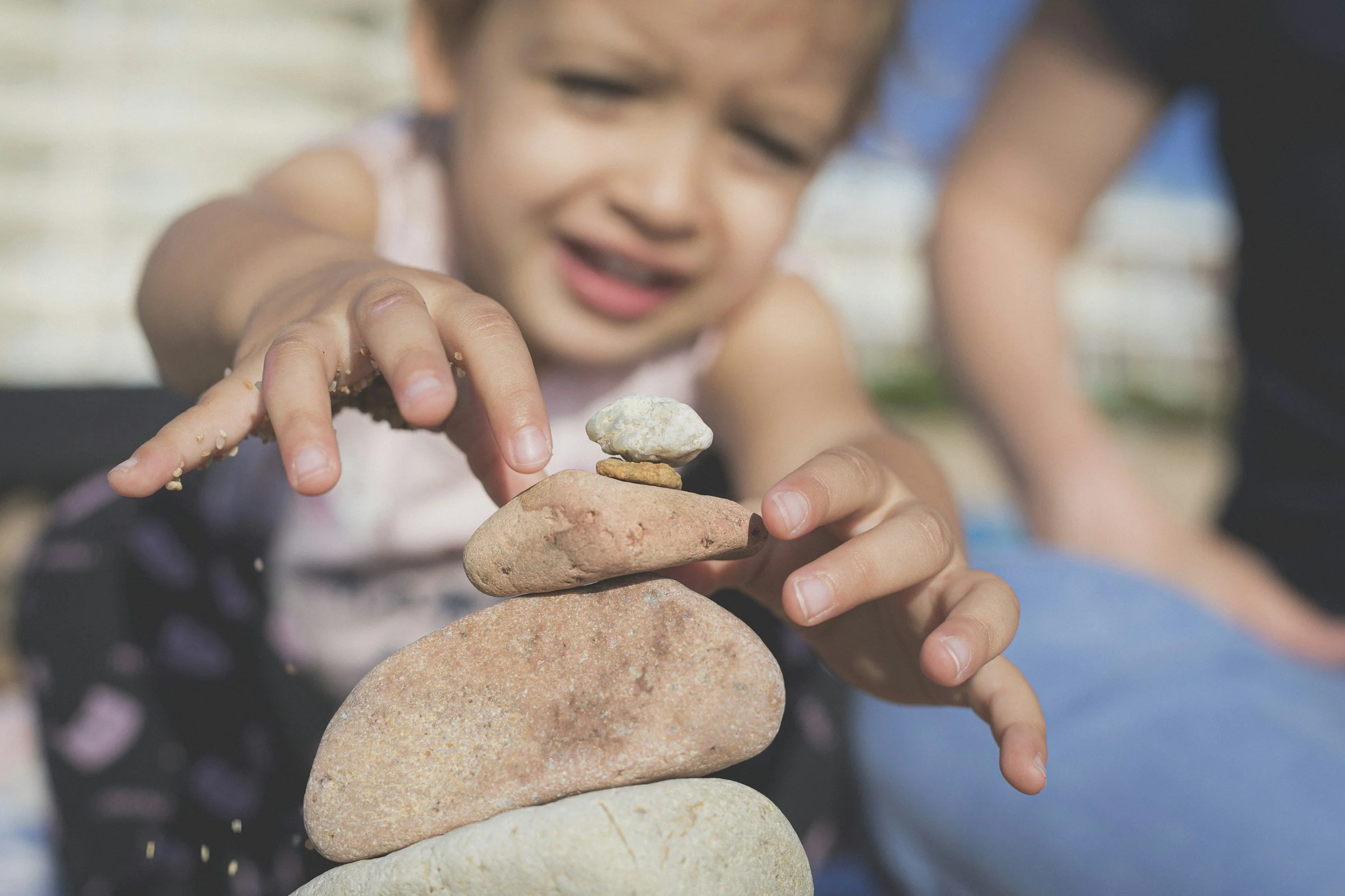 Child stacking stones outdoors, with another child visible in the background.