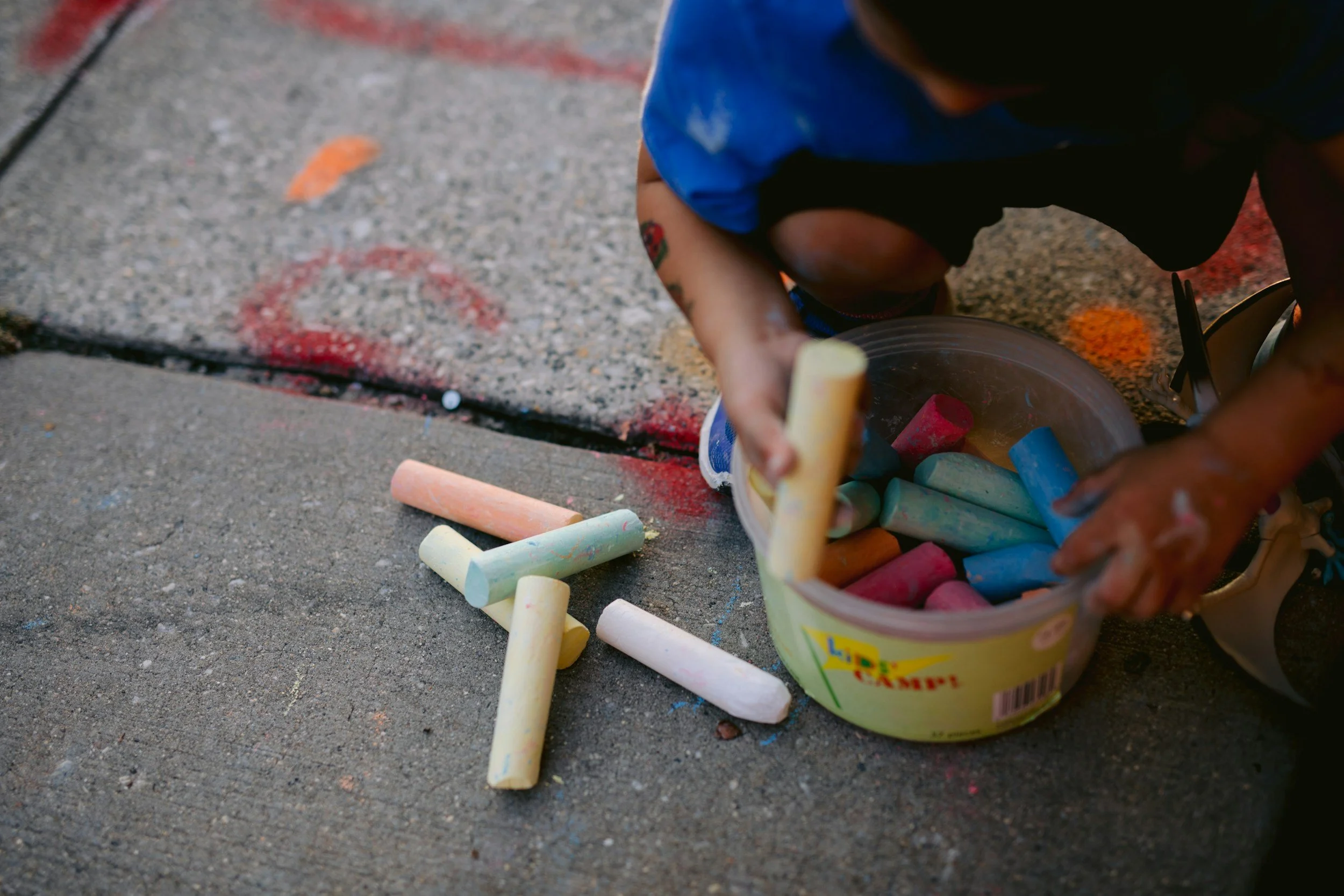 A child crouching on a sidewalk, holding a piece of chalk, with a container of colorful chalks nearby, and chalks scattered on the ground.