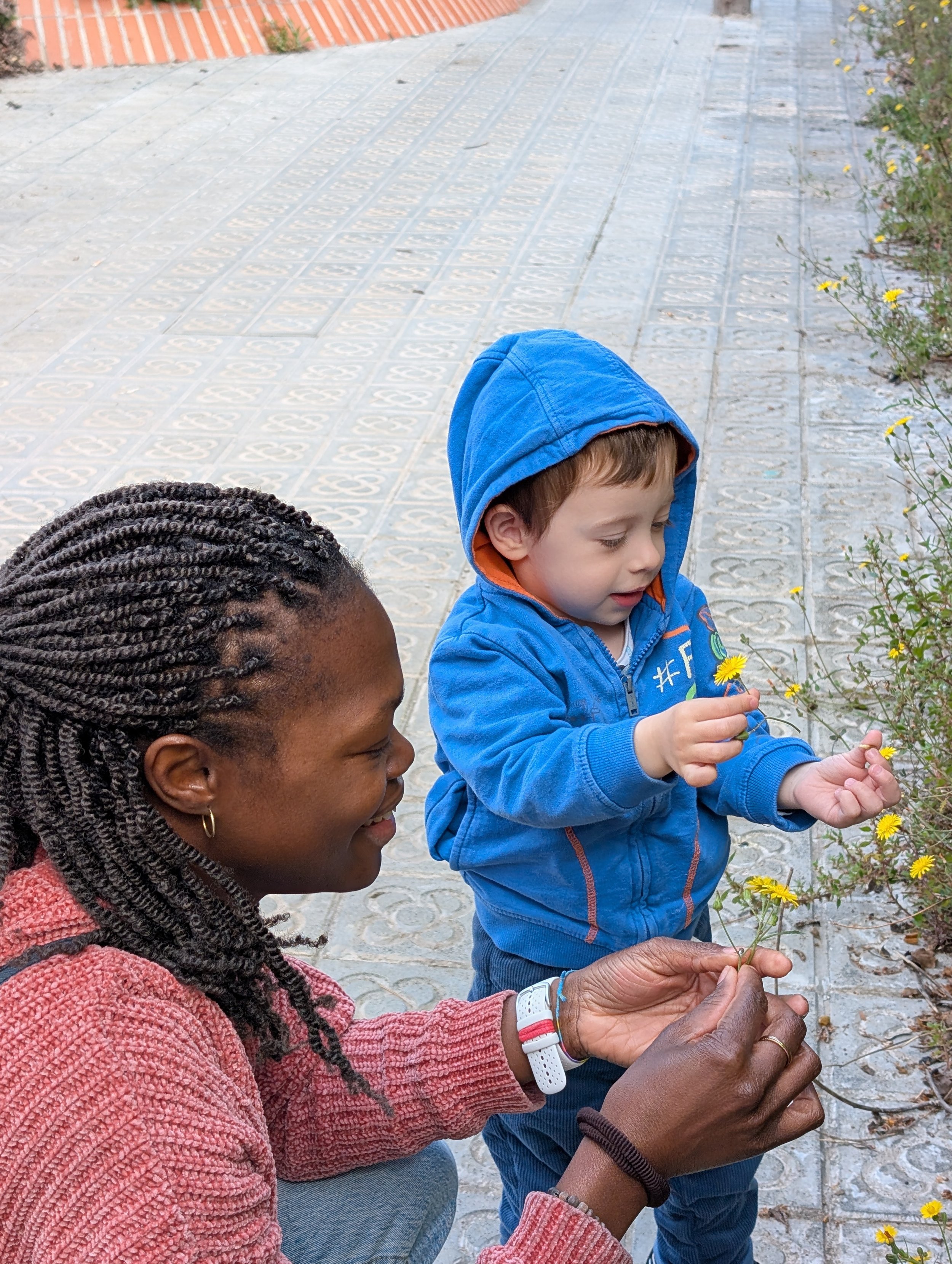 An adult woman with braided hair and a young boy with a hooded jacket picking yellow flowers together outside on a paved walkway.