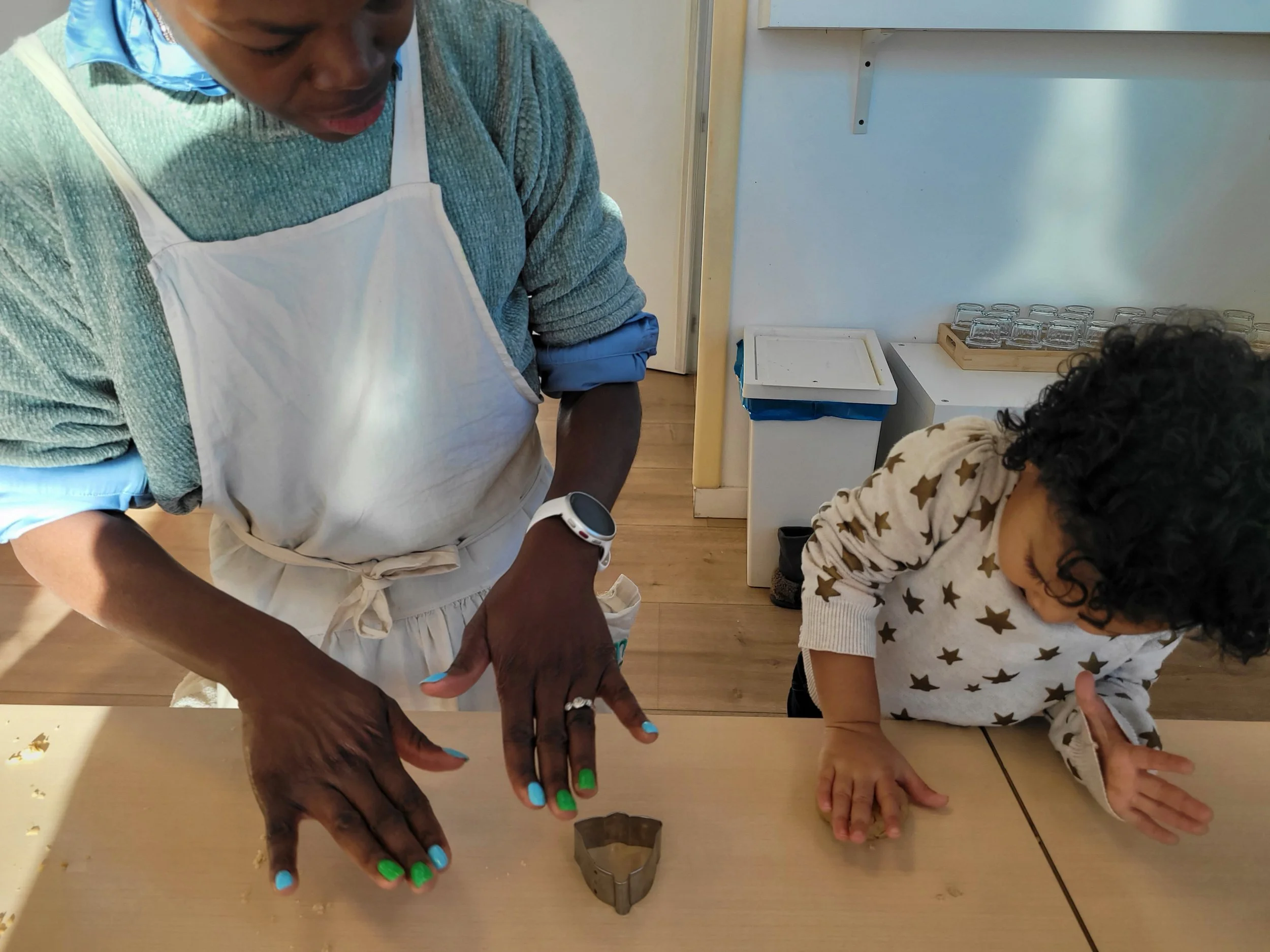 A person with multiple rings on fingers and colorful nail polish is using a cookie cutter to cut dough on a table, while a young child with curly hair and star-patterned sweatshirt looks on and points.