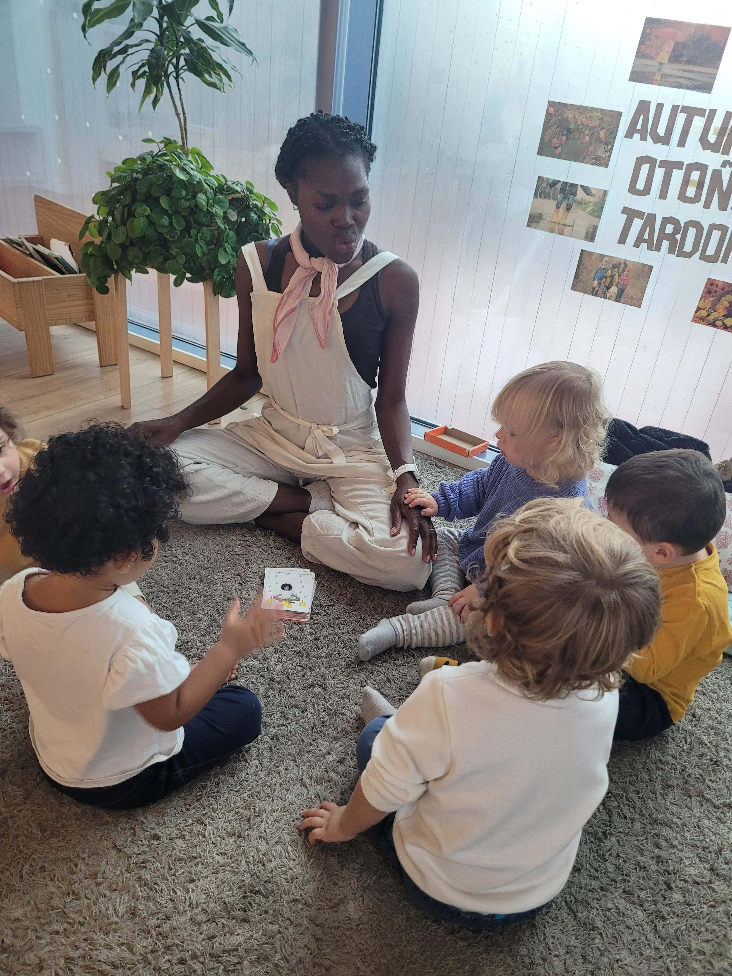 A woman sitting cross-legged on a rug, engaging with five children in a classroom or activity room, with a poster on the window and potted plant in the background.