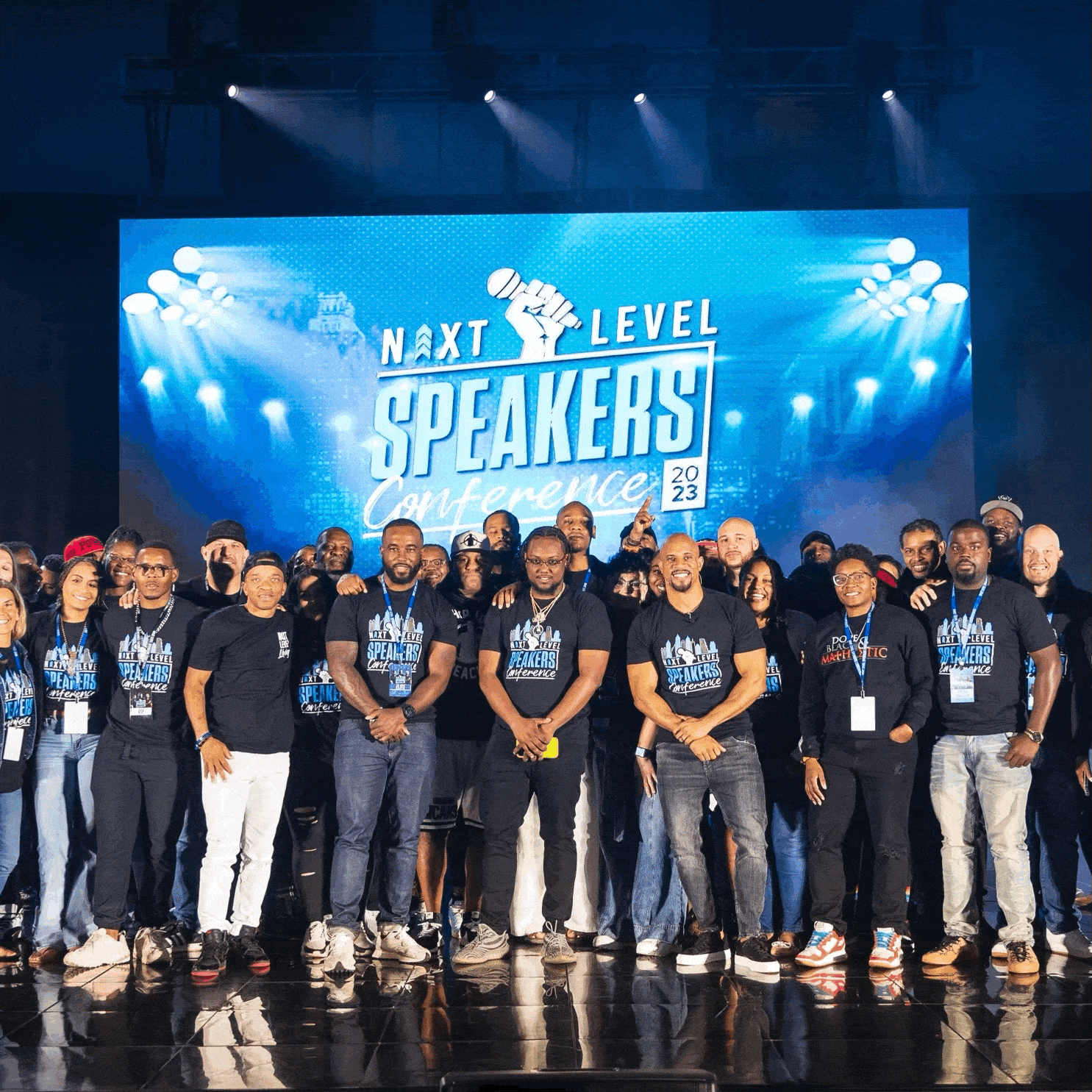 A group of people standing on a stage at the Next Level Speakers Conference 2023, with a large screen behind them displaying the conference name and logo.