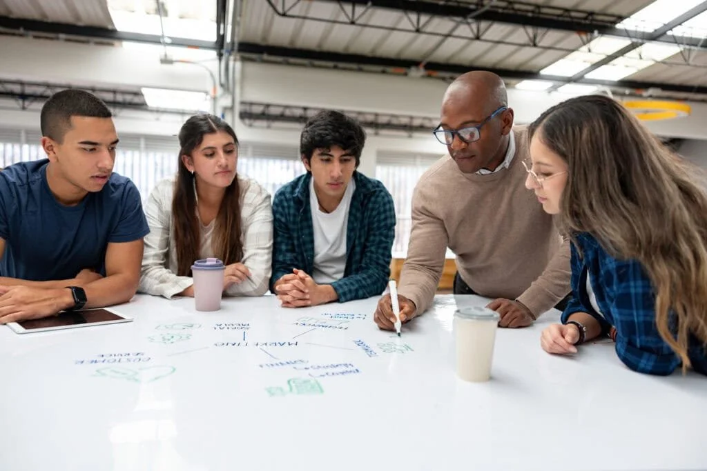 A group of five young people and a middle-aged man are gathered around a table in a modern office or coworking space, engaged in a discussion or brainstorming session, with some notes and coffee cups on the table.