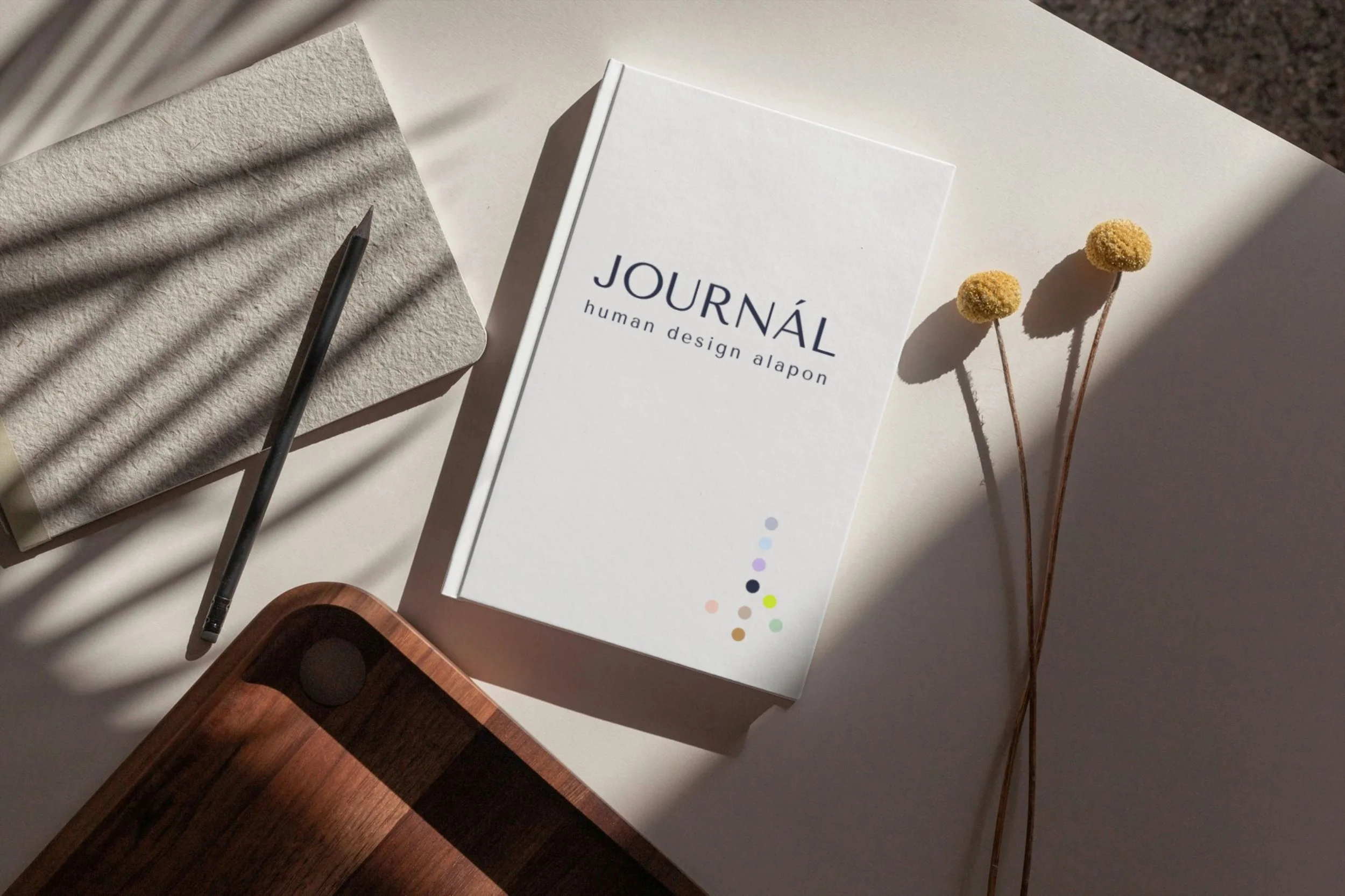 A white notebook titled 'JOURNÁL' with the subtitle 'human design alapon,' a grey textured notepad with a pen on top, two dried yellow flowers, and a wooden object, all arranged on a white surface with sunlight and shadows.