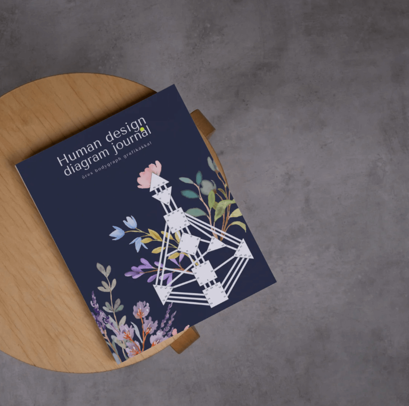 A magazine titled Human Design Diagram Journal resting on a round wooden surface, with a floral illustration on the cover, against a concrete floor background.