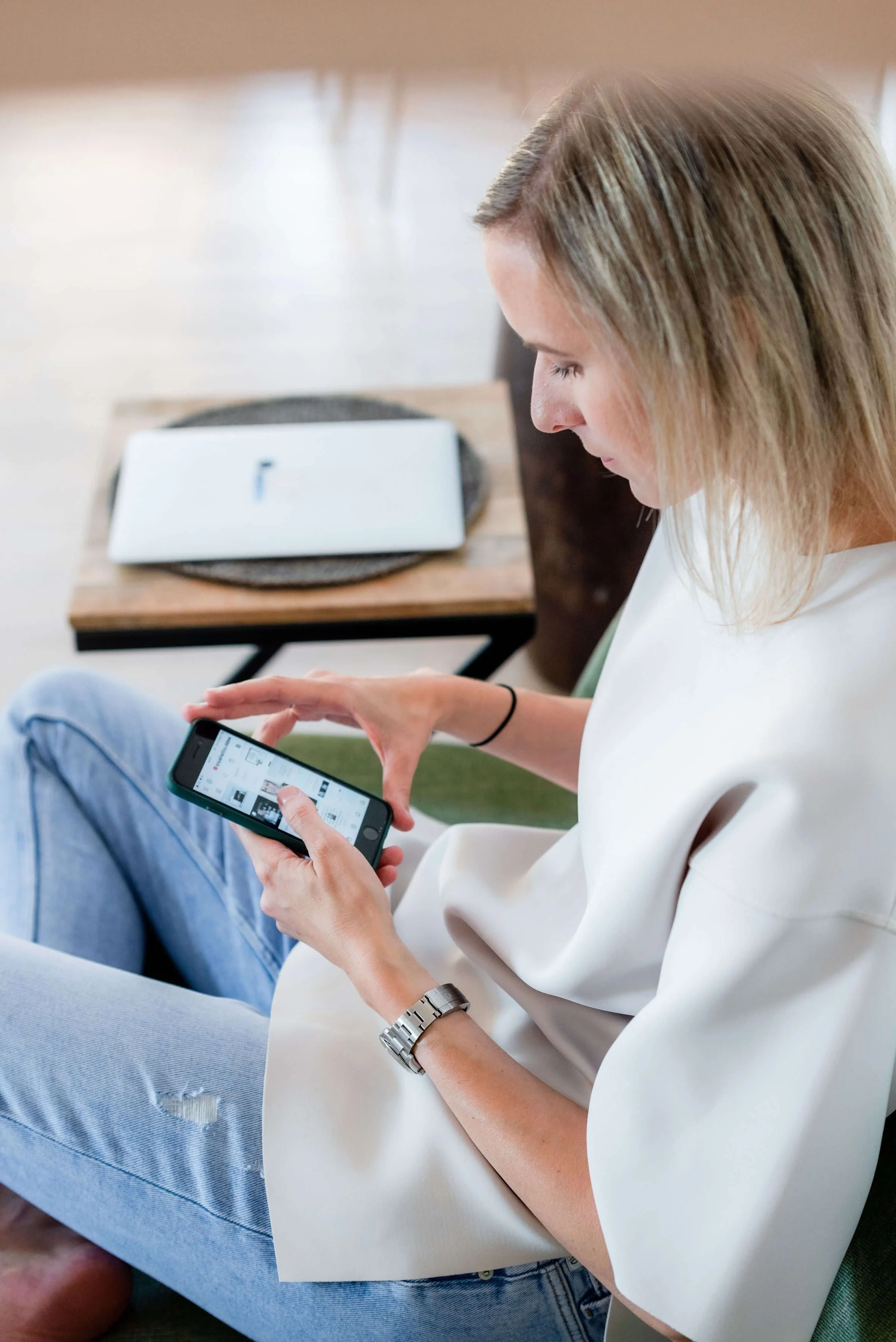 Young woman with blonde hair and a white shirt sitting on green chair, looking at her smartphone, with a watch on her left wrist, in a casual indoor setting.