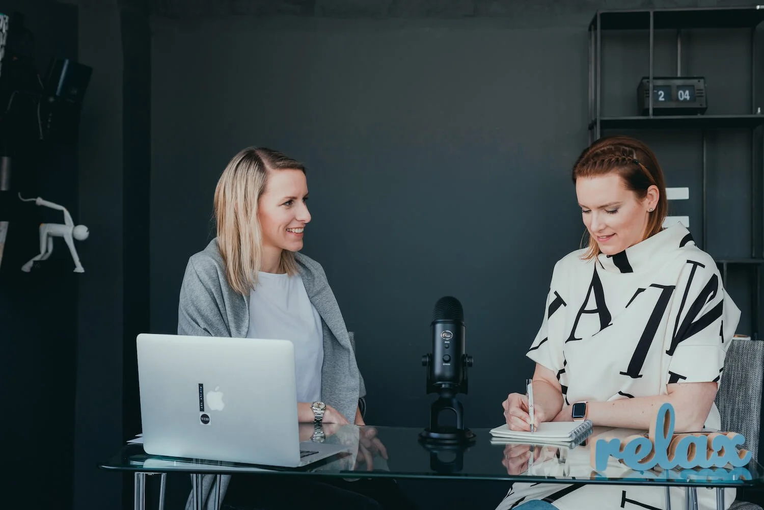 Two women sitting at a desk with a laptop and microphone, engaged in conversation. One is smiling, the other is writing in a notebook. The setting appears to be a podcast or recording studio.