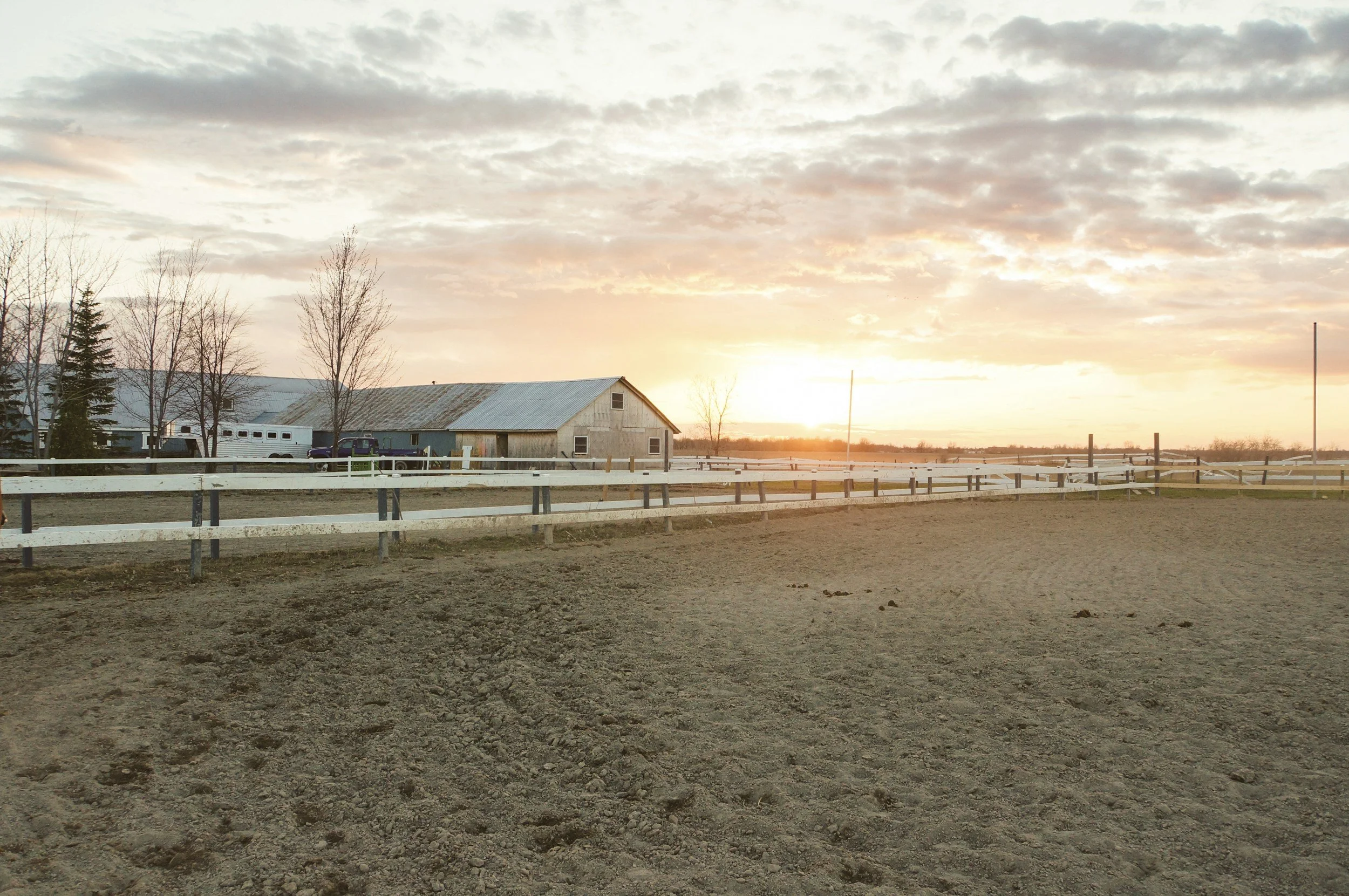 Blick auf einen Pferdehof bei Sonnenuntergang, mit Scheune, Zaun und freiem Feld.