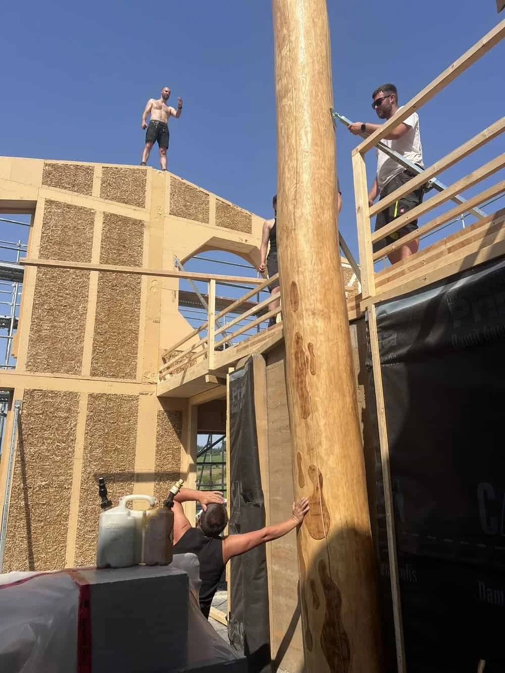 Menschen bauen ein Holzhaus, zwei befinden sich auf dem Obergeschoss, einer hält einen Gegenstand, während eine Person an einer großen Holzsäule arbeitet.