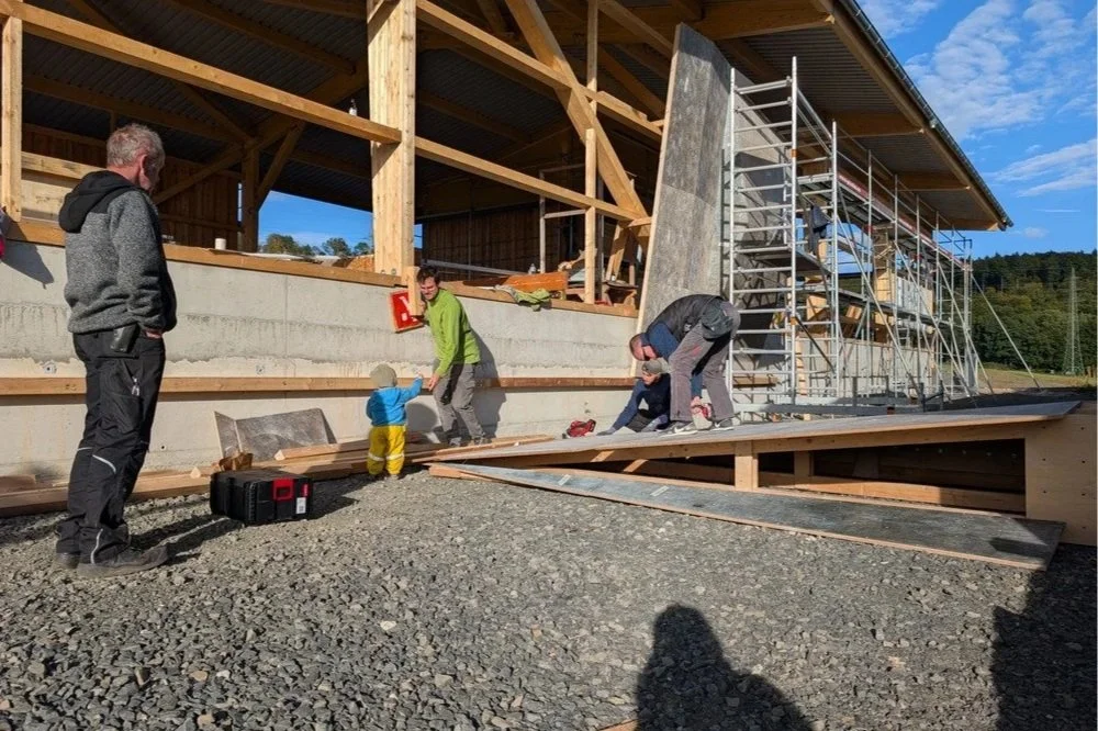 Menschen bauen ein Holzhaus auf einer Baustelle, mit Gerüsten und Baumaterialen im Freien bei Sonnenwetter.