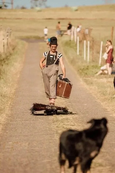 Eine Frau im Vintage-Stil fährt auf einem Roller auf einer Landstraße, während ein Hund im Vordergrund läuft. Im Hintergrund sind mehrere Menschen an einer Zaunlinie und auf einer Wiese zu sehen.