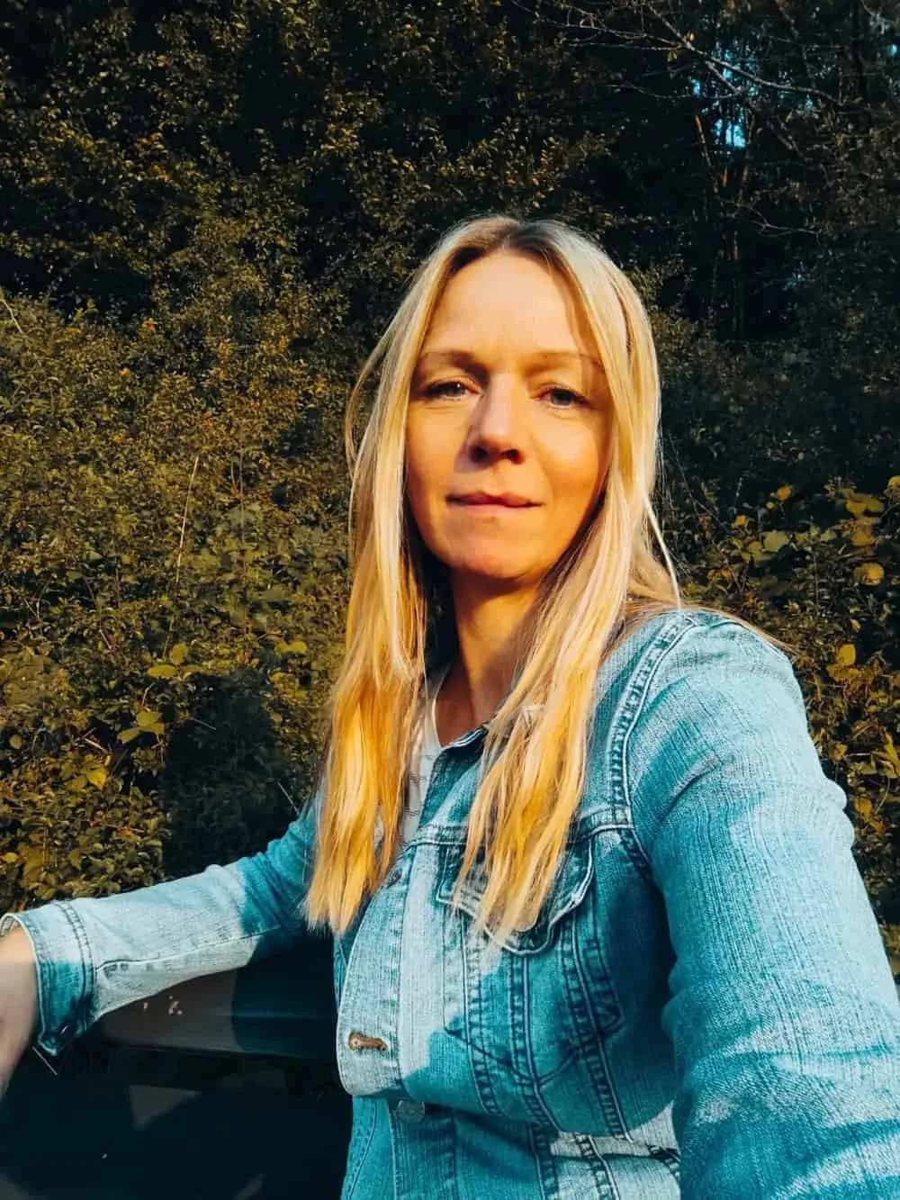 Junge Frau mit blonden Haaren in Jeansjacke vor einem bewaldeten Hintergrund bei Sonnenuntergang.