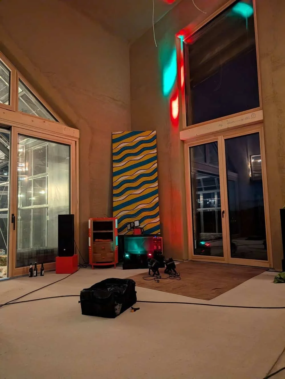 Ein unvollendeter Raum mit großen Fensterfronten, bunten Wanddekorationen und technischer Ausrüstung für Licht- und Tonaufnahmen, inklusive Kabel, Lautsprecher und einem Werkzeugkoffer.