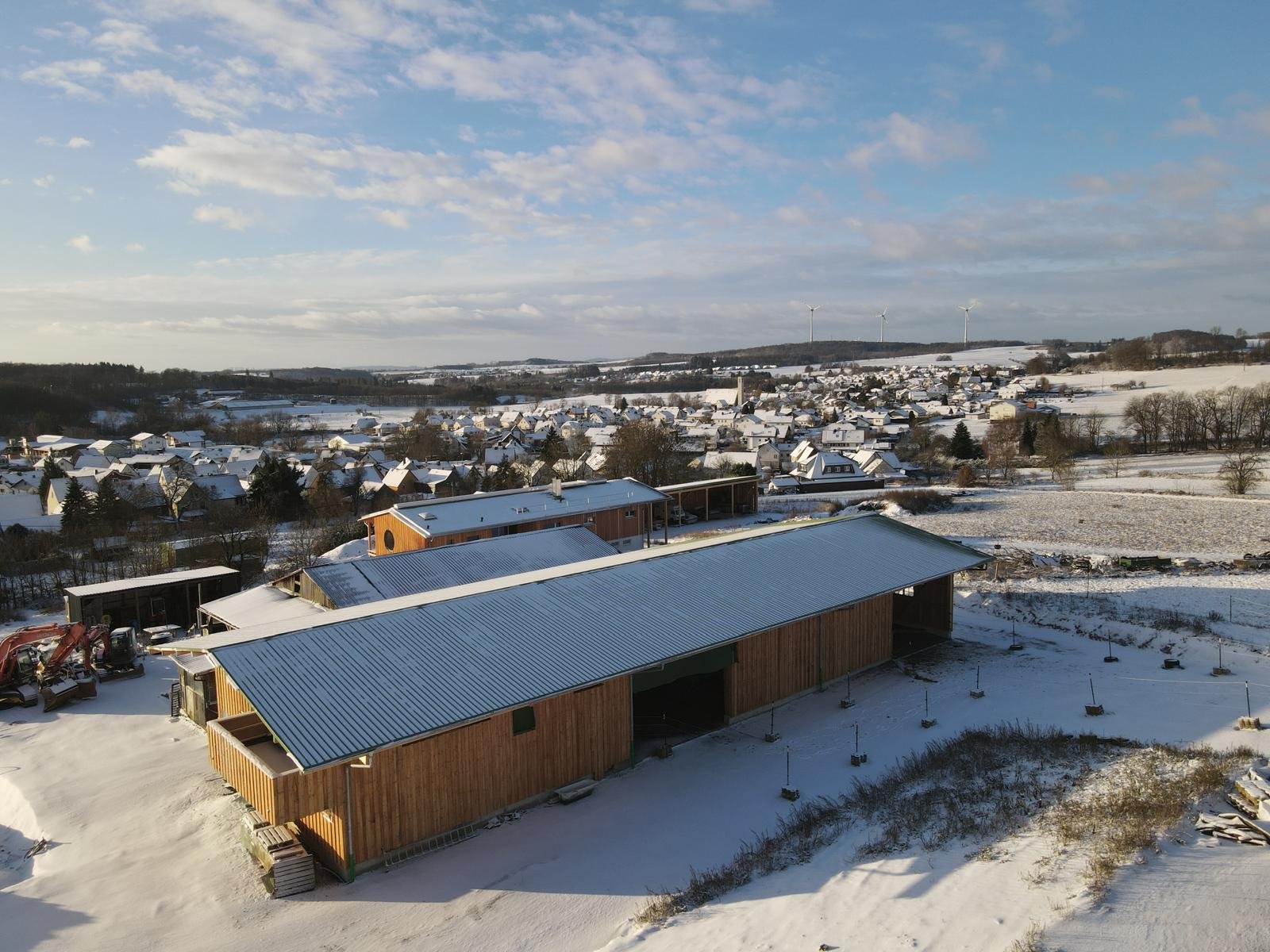 Blick auf eine verschneite ländliche Landschaft mit Gebäuden, Bäumen, Windrädern im Hintergrund und einem blauen Himmel mit Wolken.