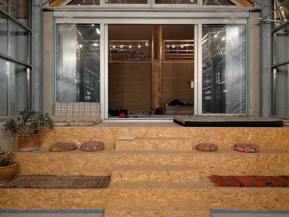 Innenraum mit Steinstufen, Sitzkissen, einer Vertiefung im Hintergrund, Holzwand und Pflanzen.