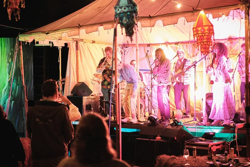 Eine Live-Band spielt auf einer kleinen Bühne unter einem Zelt bei Nacht, mit mehreren Musikern, die Instrumente spielen, und einem Publikum, das zuschaut.