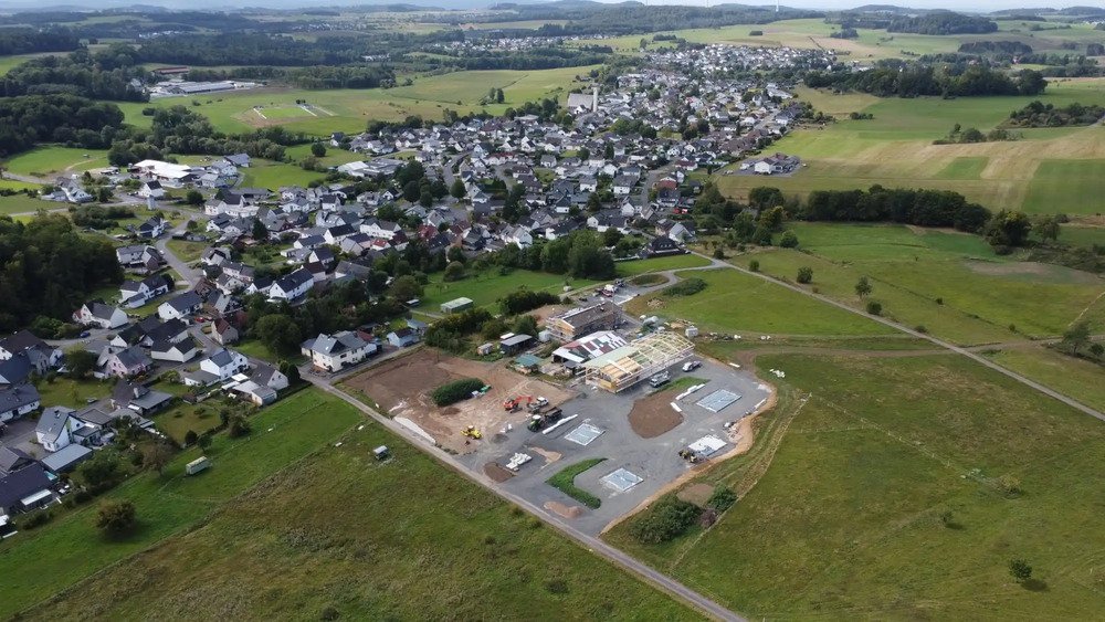 Luftaufnahme eines Dorfes mit Baustelle im Vordergrund, umgeben von Feldern und Wäldern.