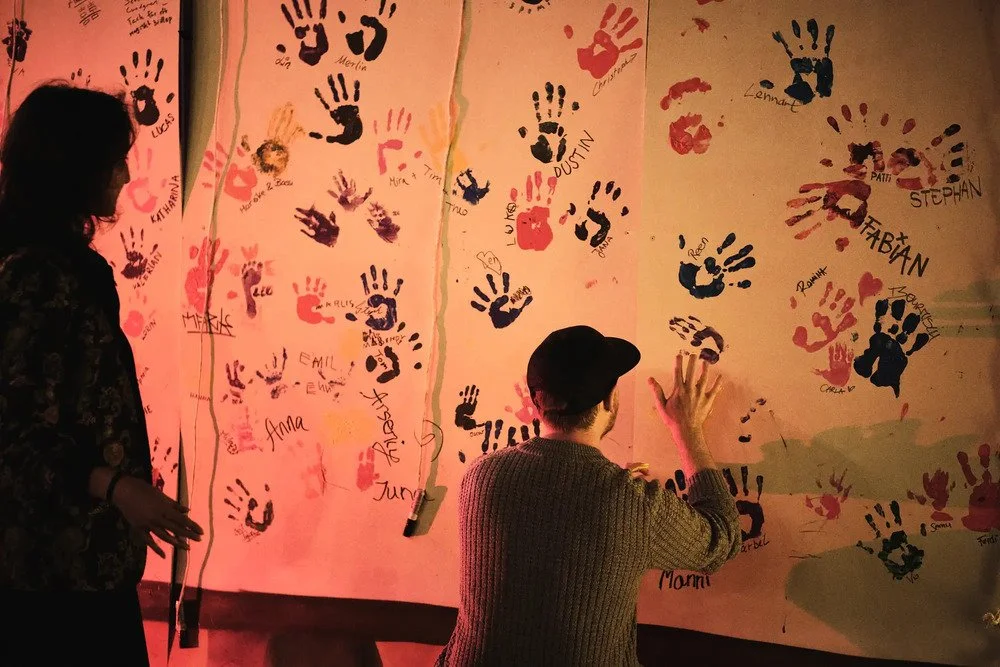 Zwei Personen malen bunte Handabdrücke und Unterschriften an eine Wand, vermutlich bei einer gemeinschaftlichen Veranstaltung.
