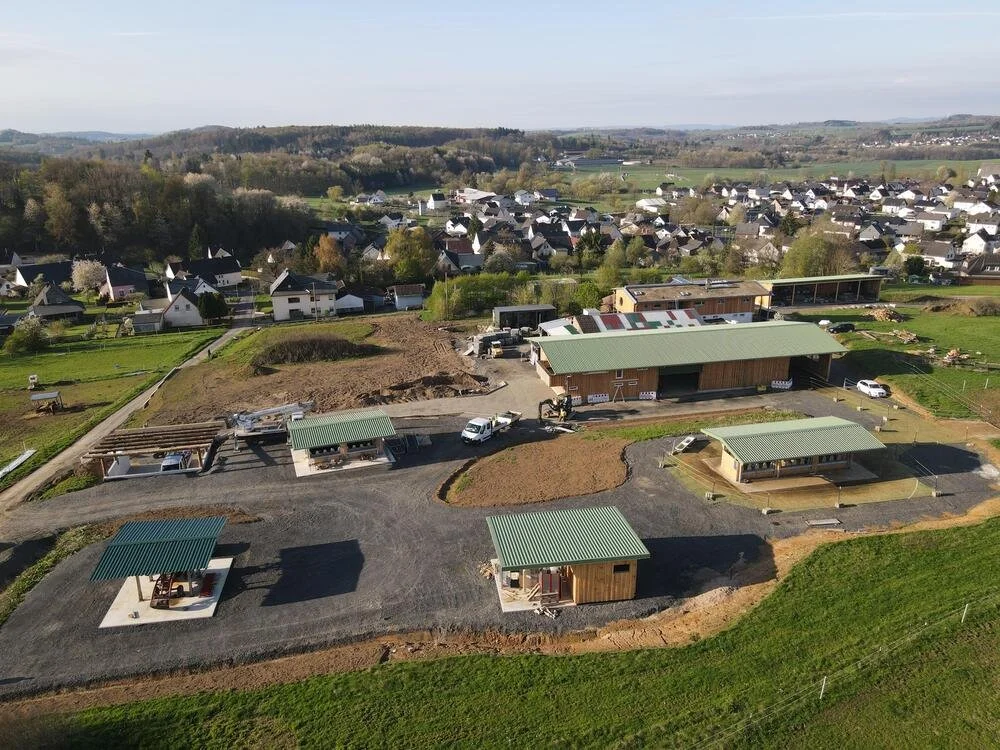 Bau von mehreren kleinen Gebäuden mit grünen Dächern auf einem Baugrundstück in einer ländlichen Umgebung, umgeben von Wiesen und einem kleinen Dorf im Hintergrund.