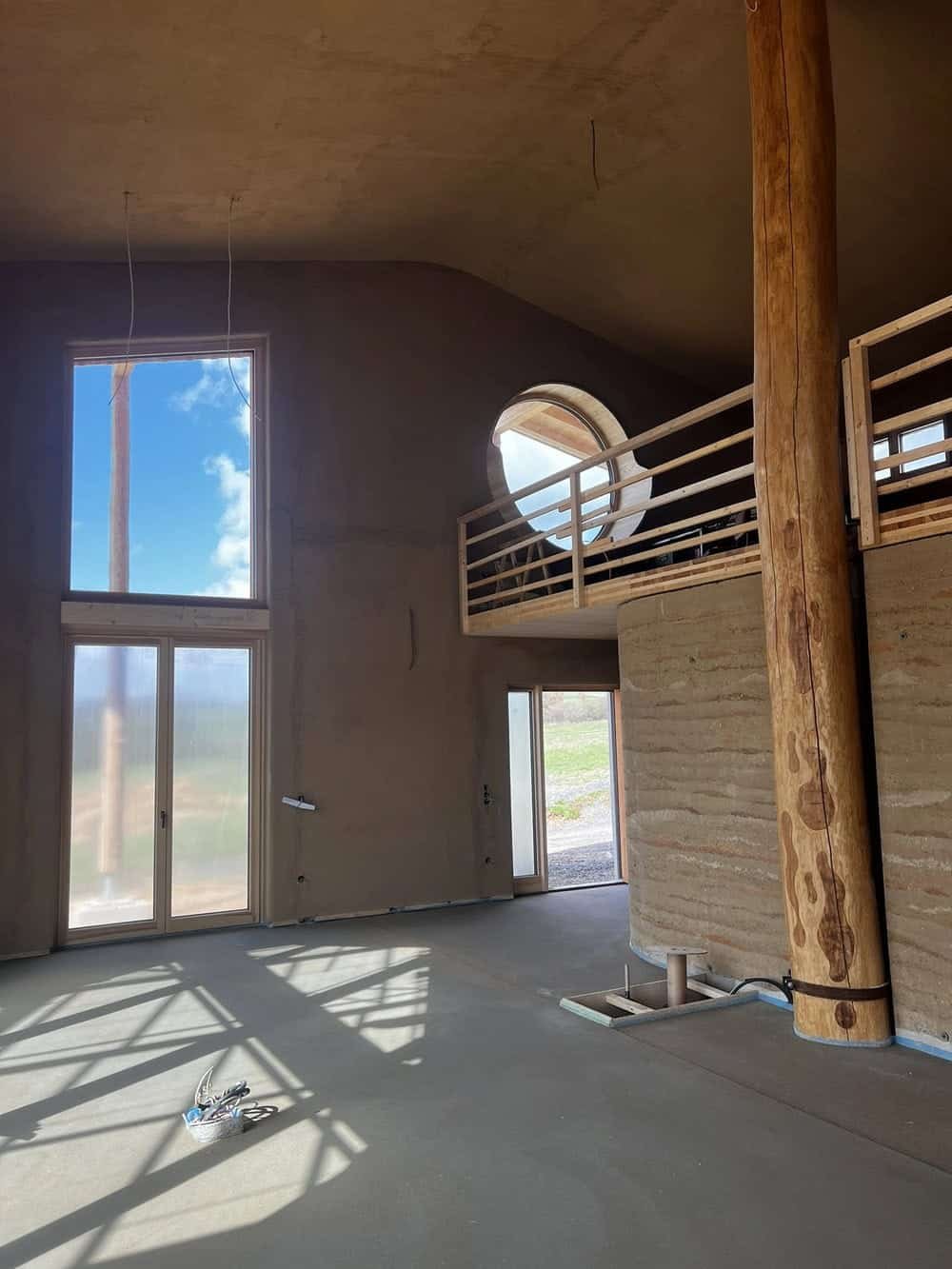 Innenansicht eines noch unfertigen Hauses mit großen Fenstern, einer Holzstütze, Stoffaltdecken und offener Fläche, mit sichtbaren Kabeln und Befestigungsschellen auf dem Boden.