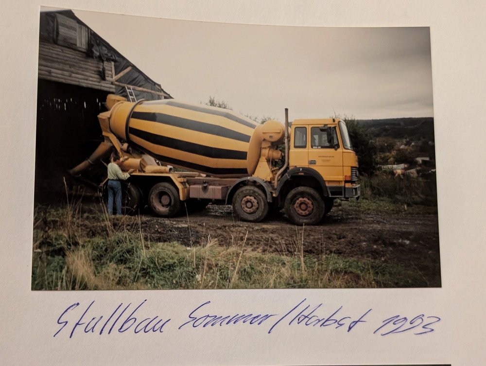 Ein gelber Baustellenwagen mit Sicherheitskegel in einer landlichen Umgebung, darunter handschriftliche Notiz in Kursive mit blauer Tinte, die den Text "Strullbau Sommer/Hasted 1993" enthält.