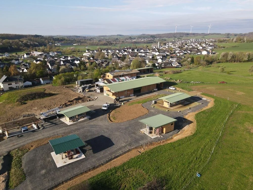 Ein Baugebiet mit mehreren neu erbauten kleinen Gebäuden und Häusern auf grünem Land, im Hintergrund eine Siedlung mit Häusern und Windkraftanlagen auf dem Horizont.