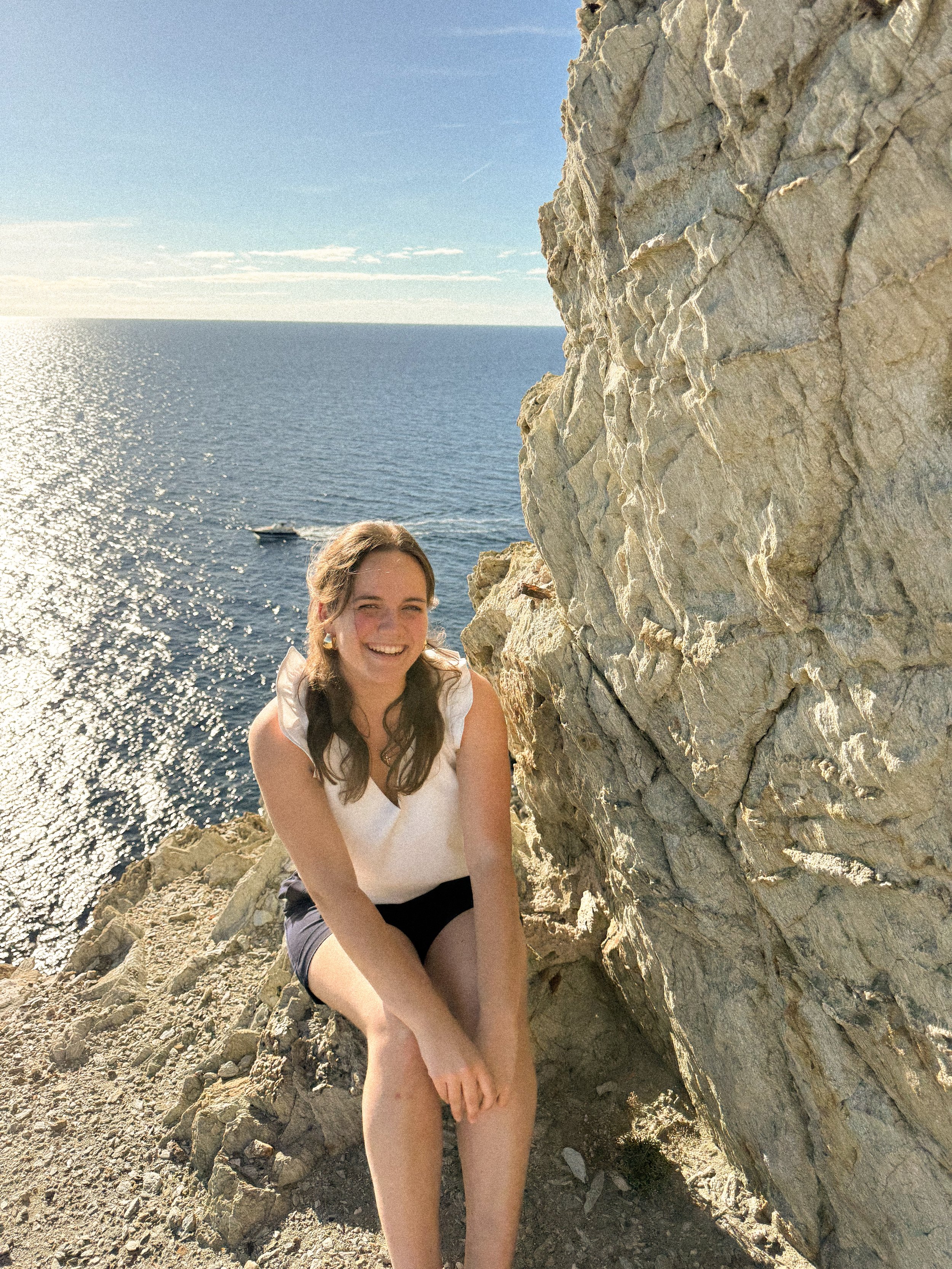 Jeune femme assise sur des rochers près de la mer, souriante, avec un bateau en arrière-plan, sous un ciel ensoleillé.