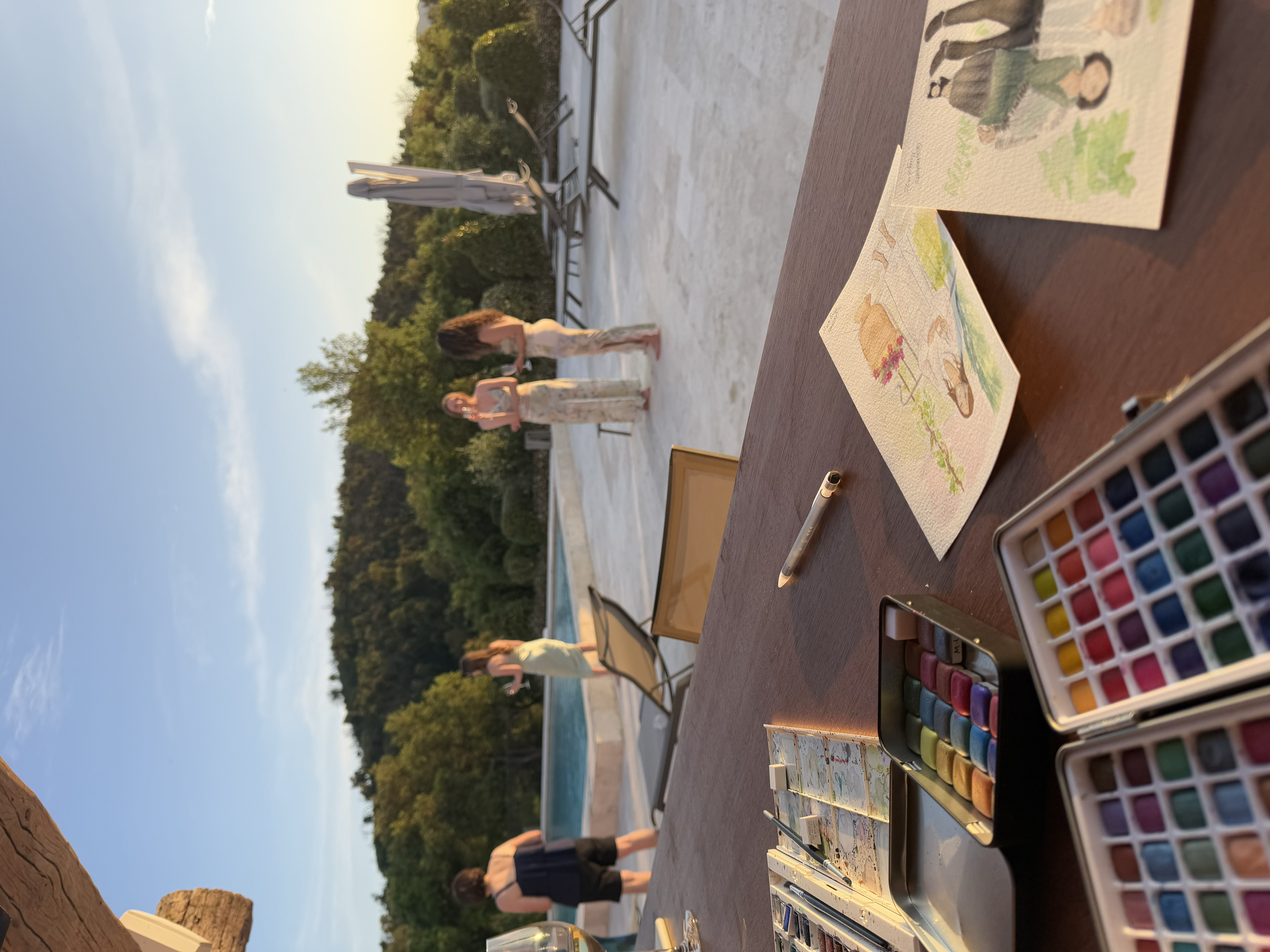 Une table de peinture avec des boîtes de couleurs aquarelles, et deux peintures avec des illustrations de personnes en plein air, vues de près. En arrière-plan, plusieurs personnes en robe debout près d'une piscine extérieure, entourées de verdure et