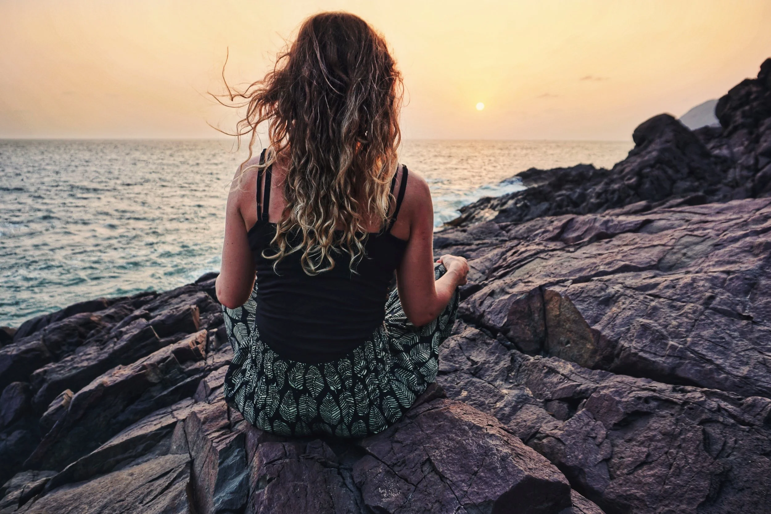 Woman with curly hair sitting on rocks by the ocean at sunset