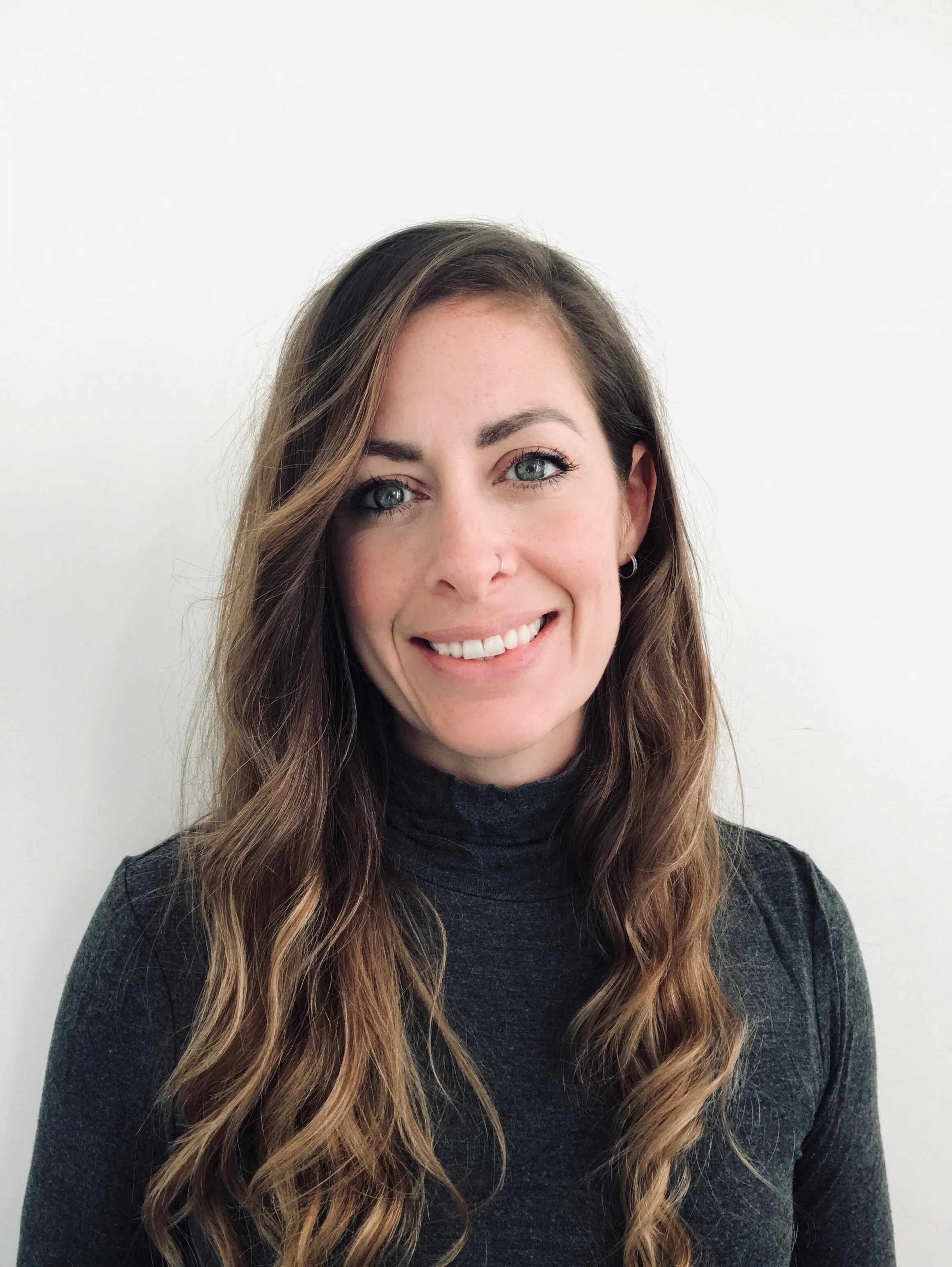 Portrait of a woman with long wavy brown hair, blue eyes, wearing a dark turtleneck, smiling in front of a plain white background.