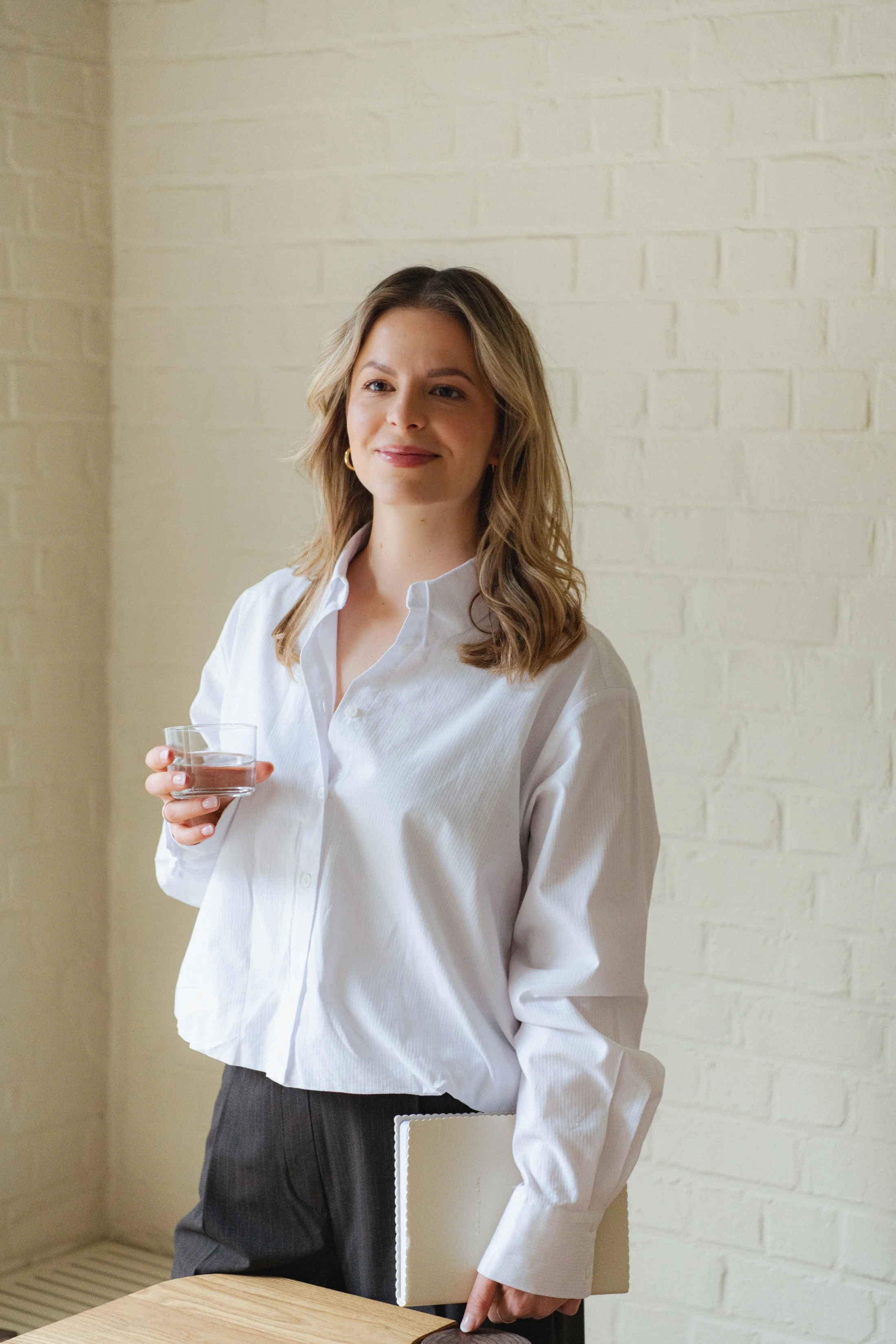Een vrouw met geluide, halflang haar, gekleed in een witte blouse en een grijze broek, die een glas water vasthoudt en een notitieboekje onder haar arm heeft, staat in een kamer met een witte bakstenen muur.