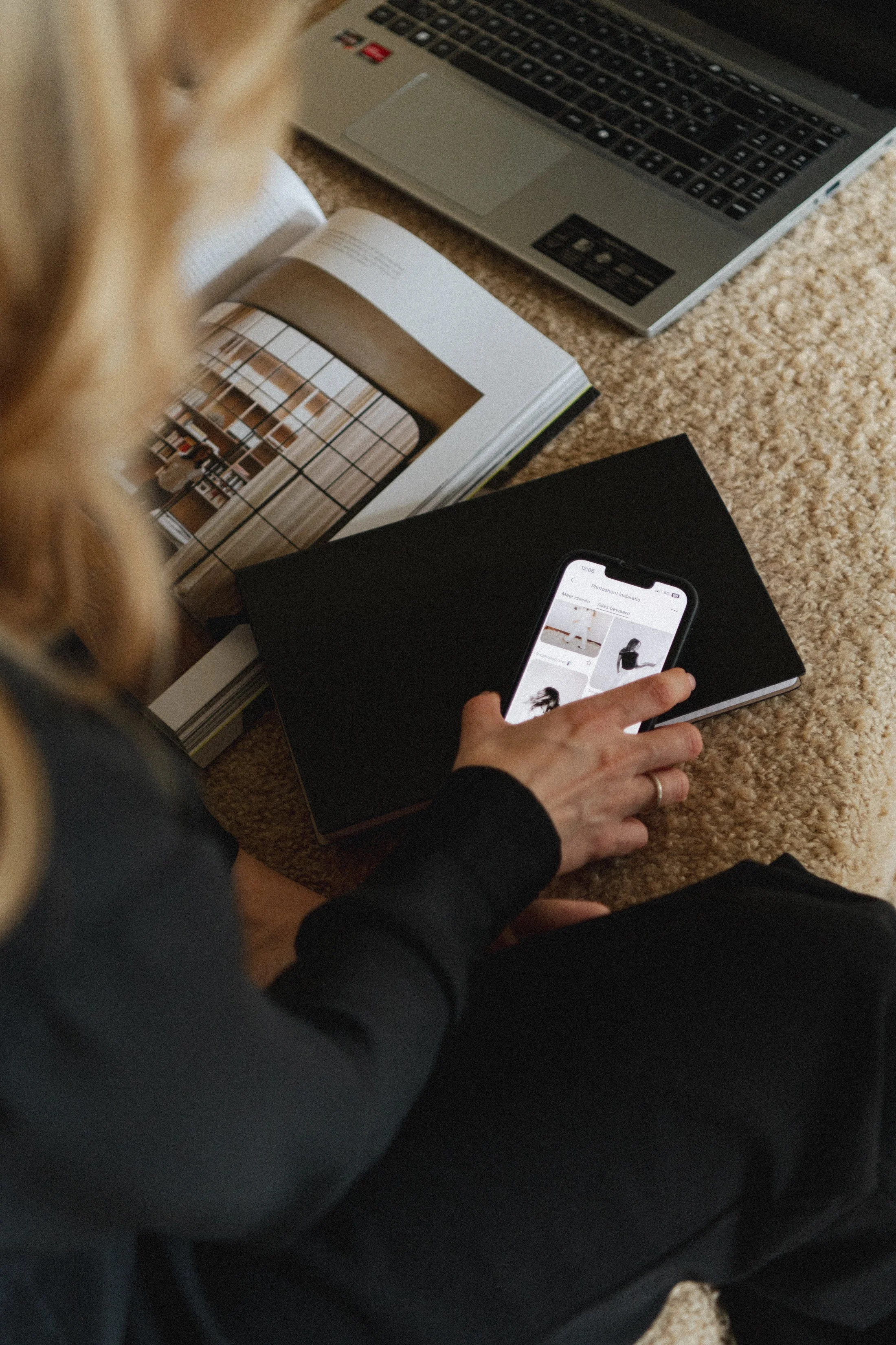 Een persoon gebruikt een smartphone en bekijkt afbeeldingen, terwijl ze naast een open laptop en een open boek met interieurfoto's op de vloer zitten.