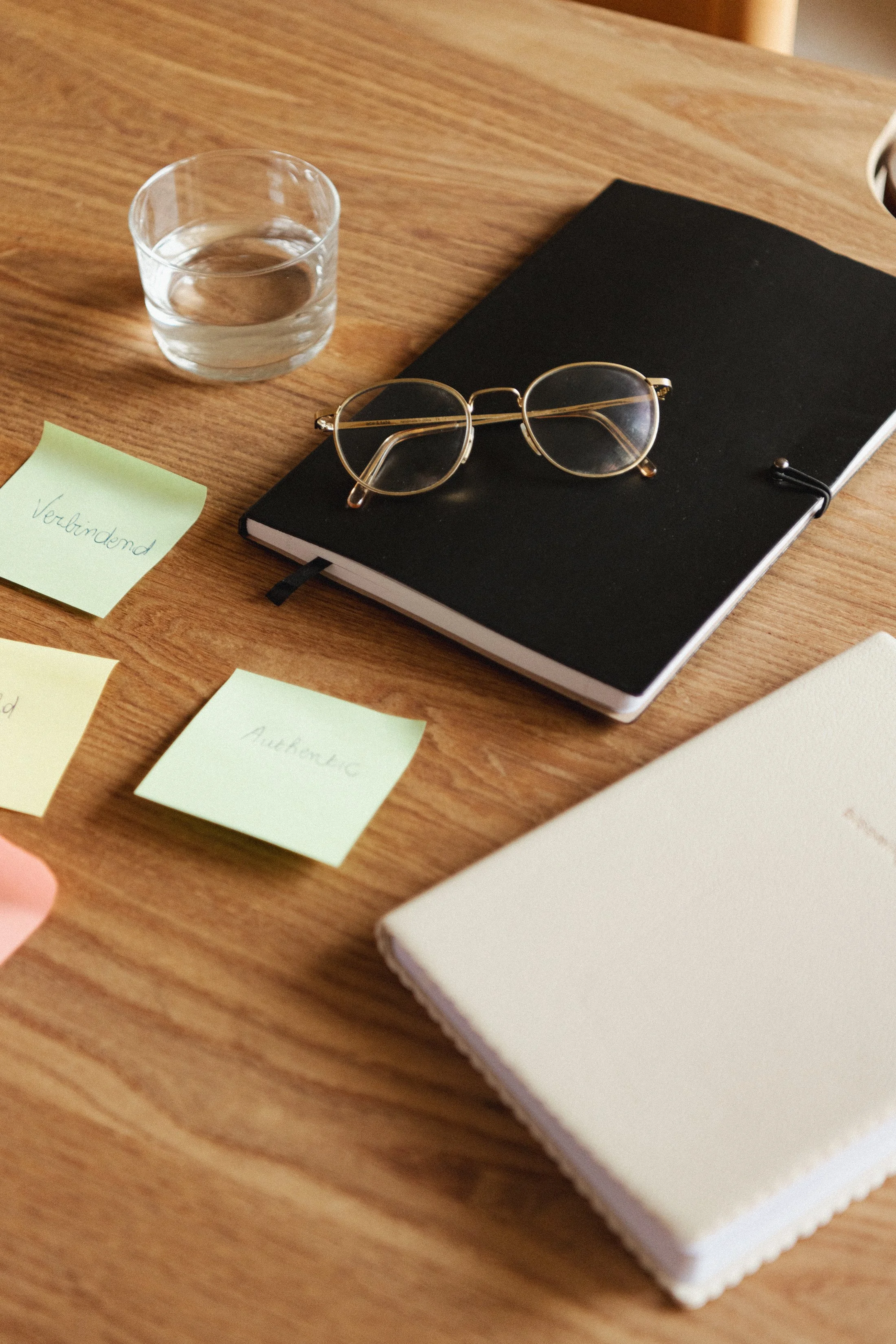 Een glas water, een zwart notitieboek en een paar zwartgekleurde bril op een houten tafel, met een post-it met 'Verbindend' geschreven.