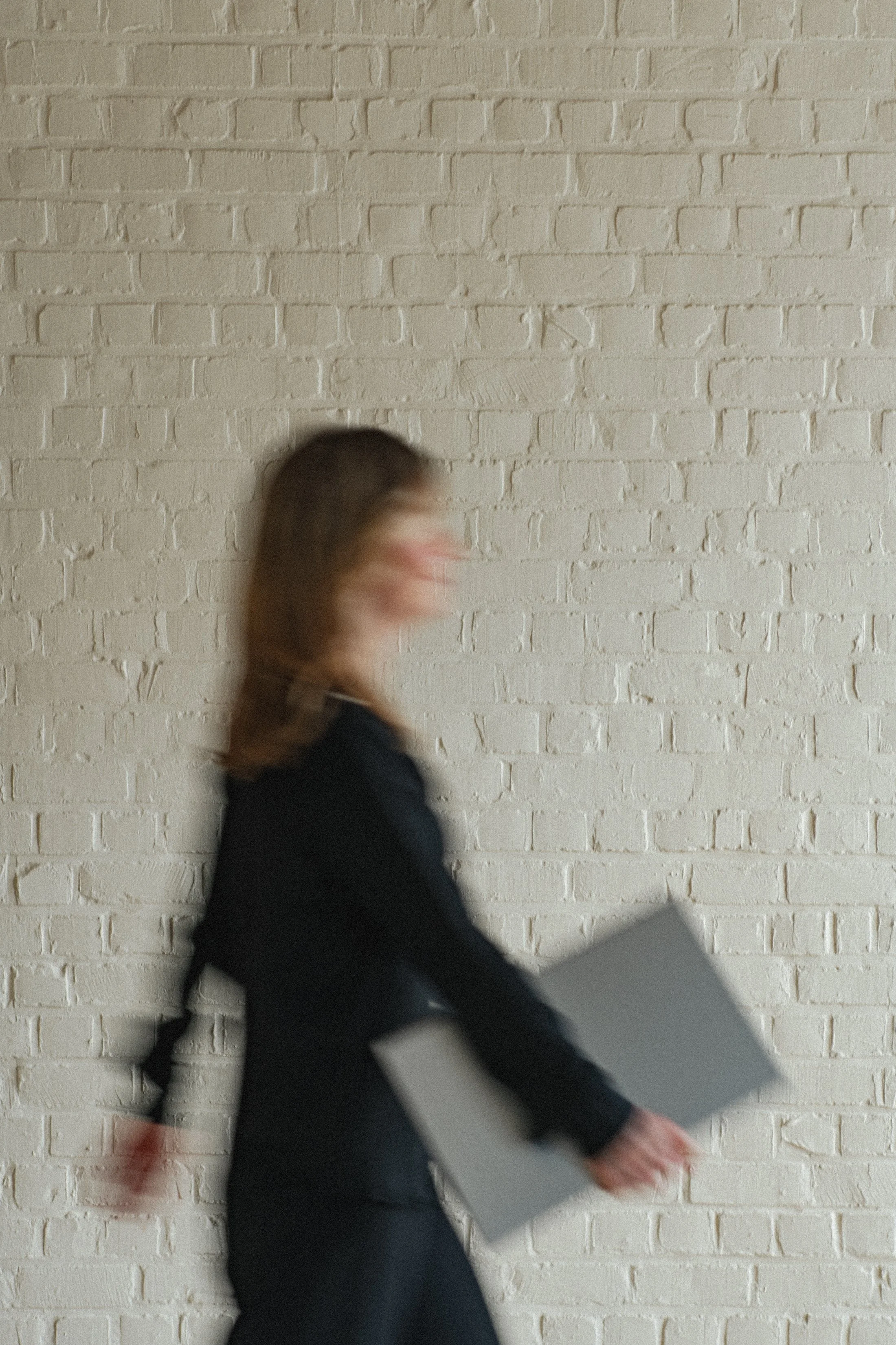 Een persoon met bruin haar in zwart kleding die loopt langs een witte bakstenen muur. De persoon lijkt te bewegen, waardoor het beeld wazig is.