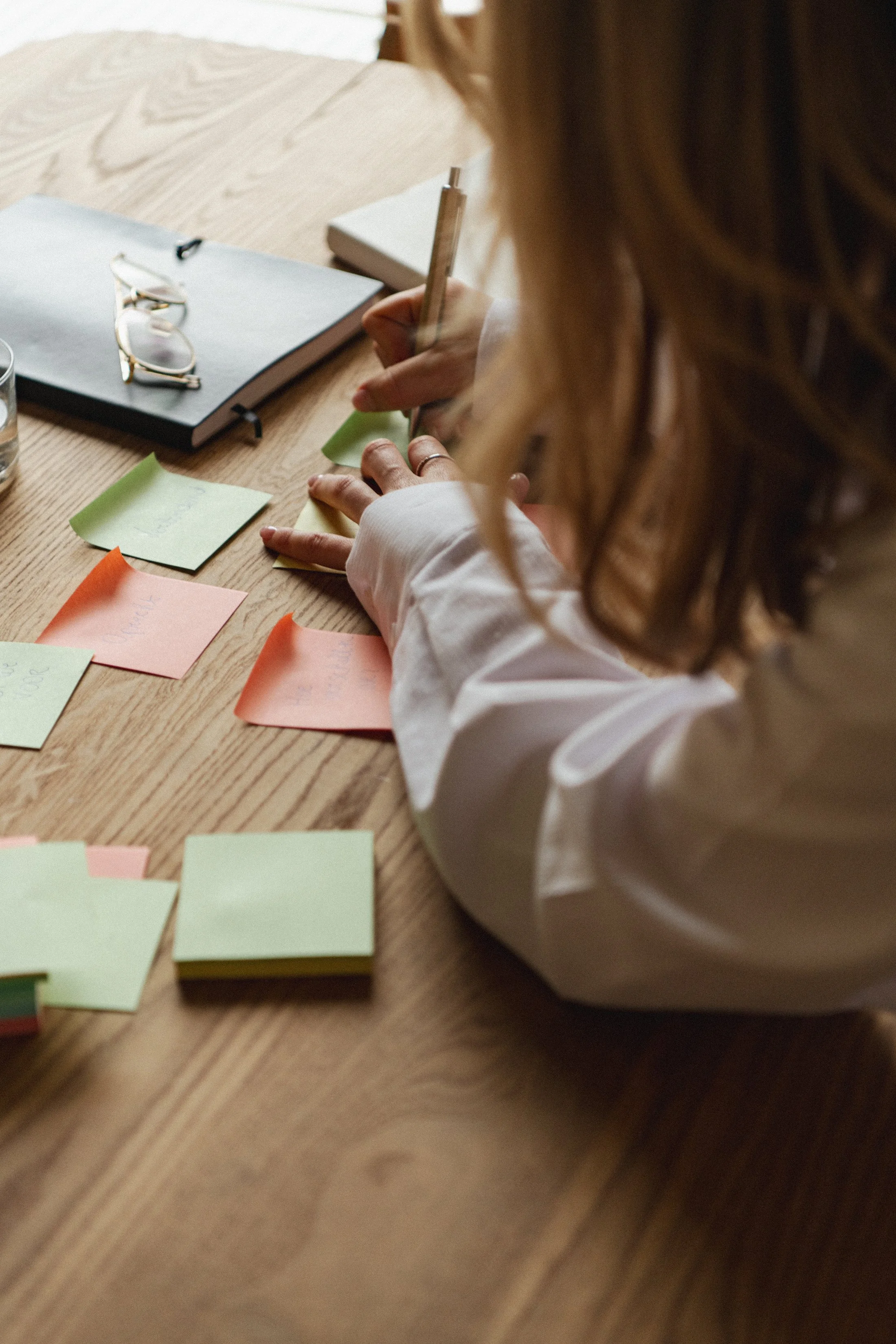 Persoon schrijft notities op gekleurde post-its op een houten tafel, met een notitieboek, een pen en een glas water op de tafel.