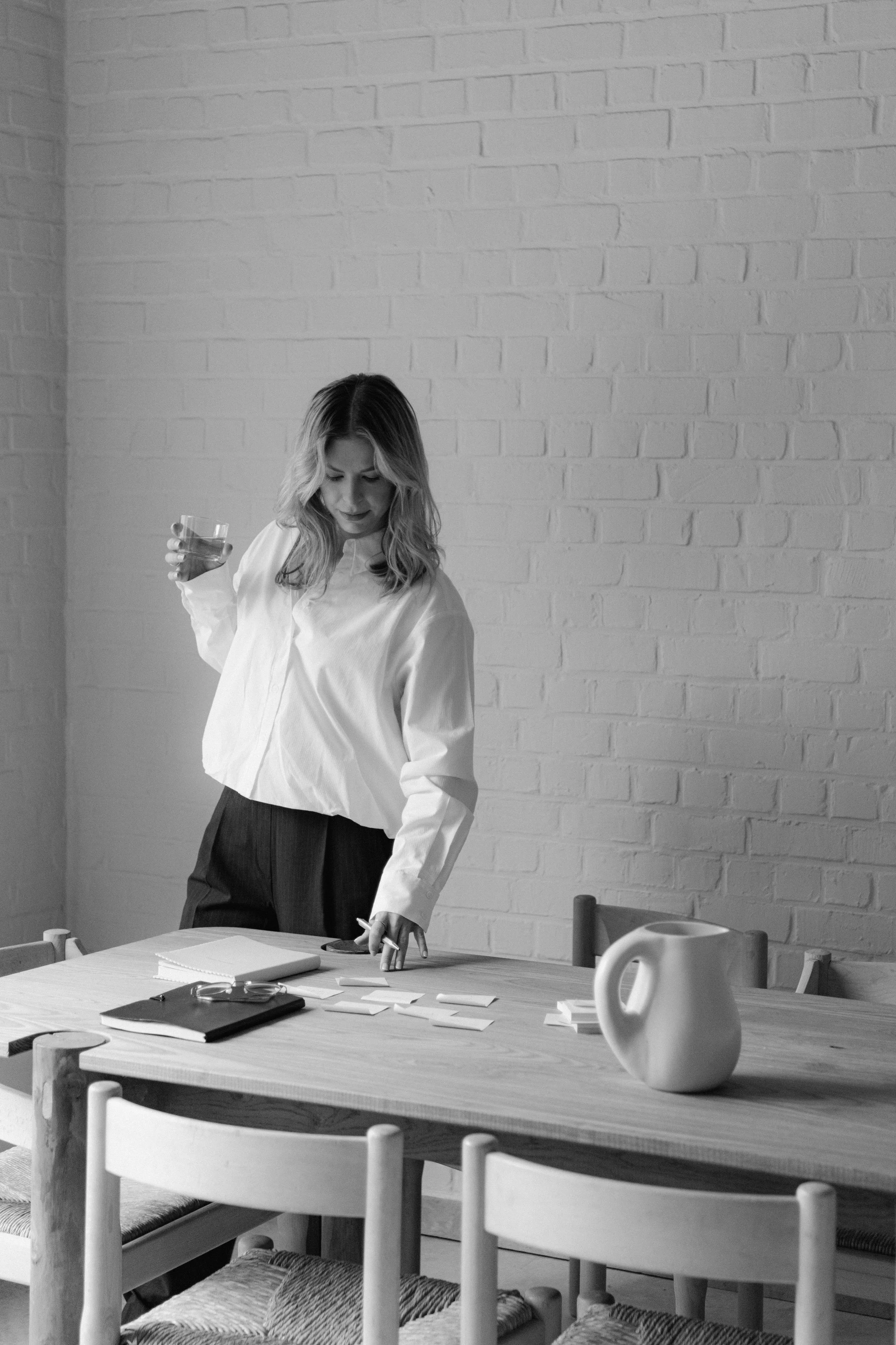 Een vrouw met lang haar draagt een witte blouse en zwarte broek. Ze staat aan een houten tafel met papieren, een notitieboek en een tablet. Ze houdt een glas water in haar rechterhand en wijst met haar linkerhand naar iets op de tafel. Op de tafel staat ook een witte vaas en een koffiekopje. Achter haar bevindt zich een witte bakstenen muur.